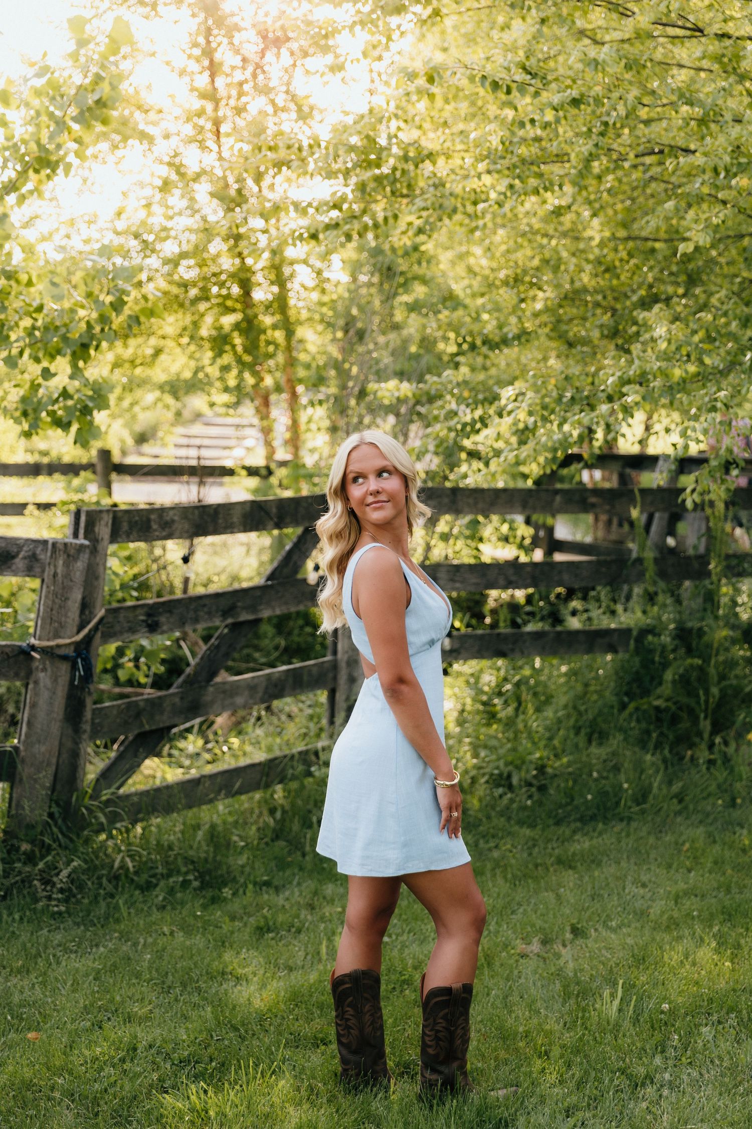 Blonde woman in white dress and cowboy boots posing by rustic wooden fence in lush green countryside.