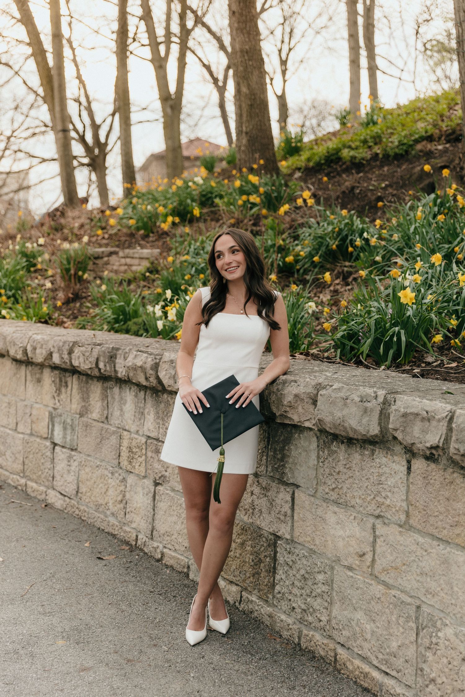 Smiling woman in white strapless dress holding diploma by stone wall with spring daffodils.