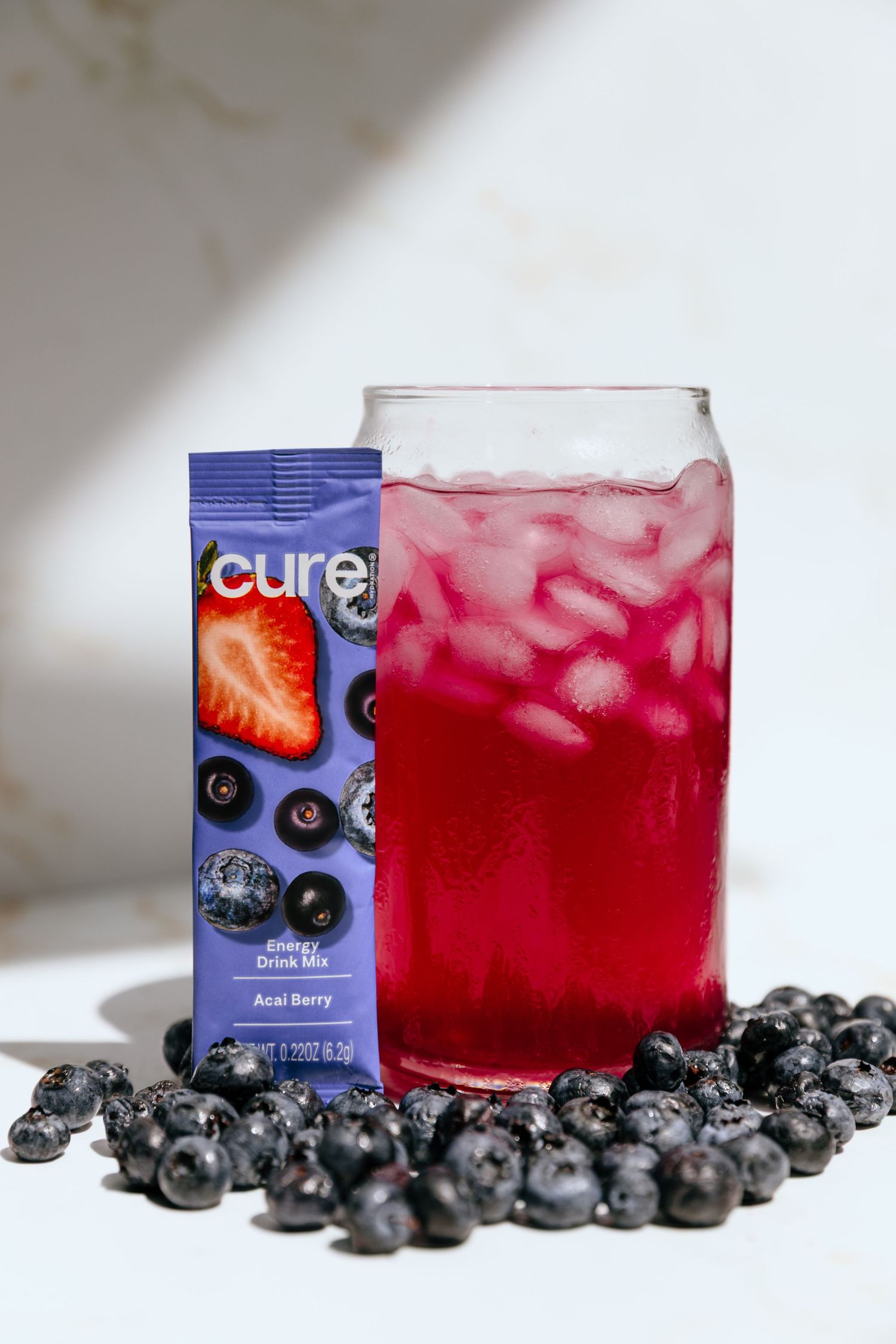 Cure hydration mix packet with fresh blueberries and pink drink in glass on white background.