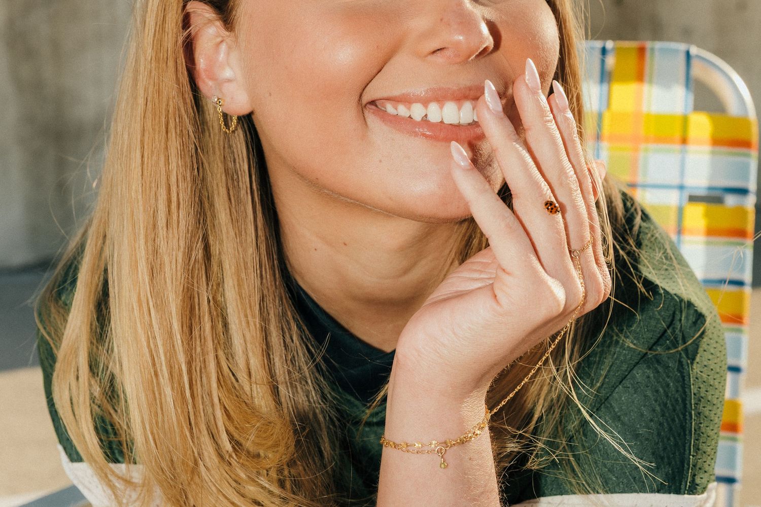 Smiling blonde woman with gold bracelet and earrings, hand near face, wearing green top, seated in colorful chair.