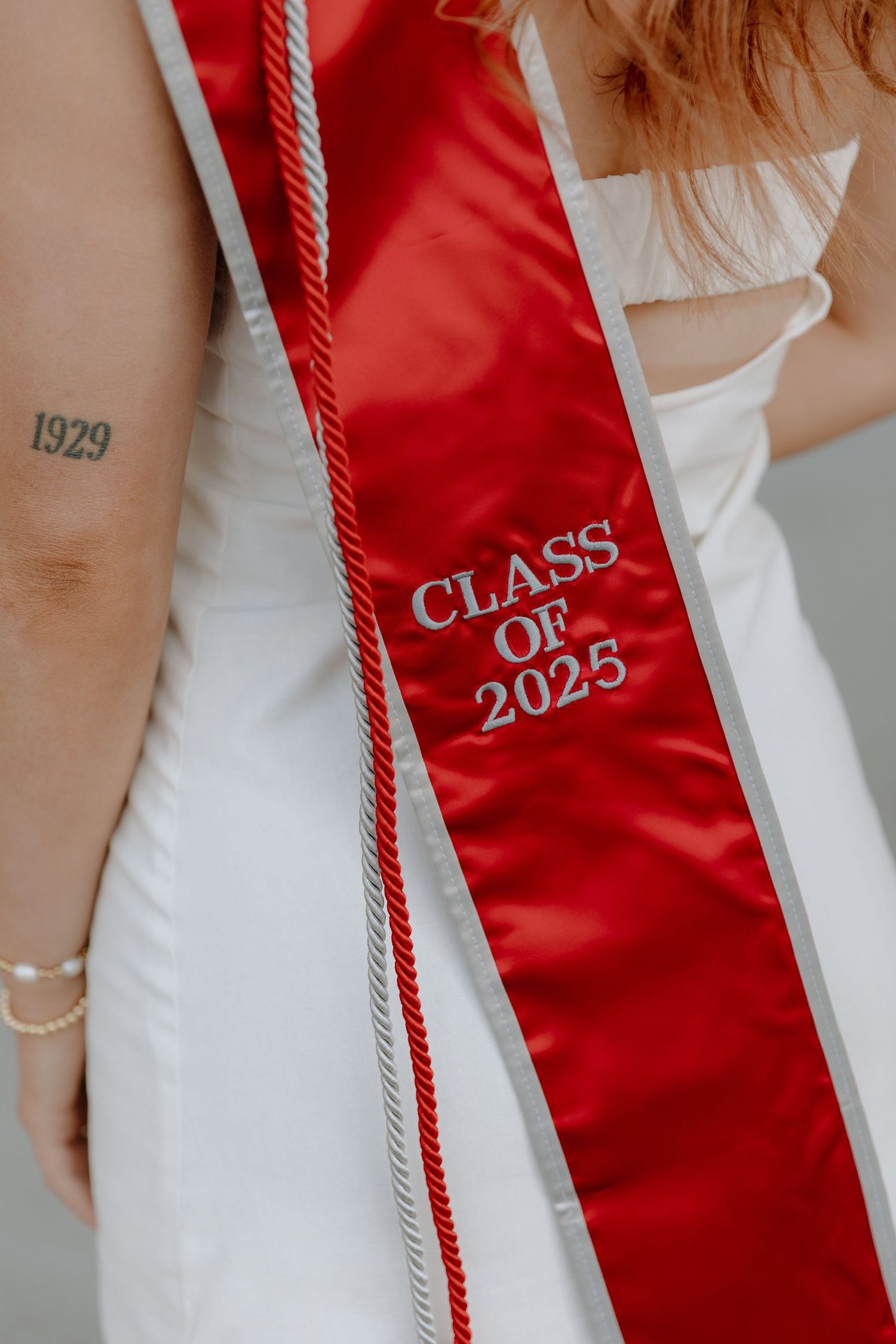 A red graduation stole with 'CLASS OF 2025' embroidered text worn over a white dress with a visible arm tattoo.
