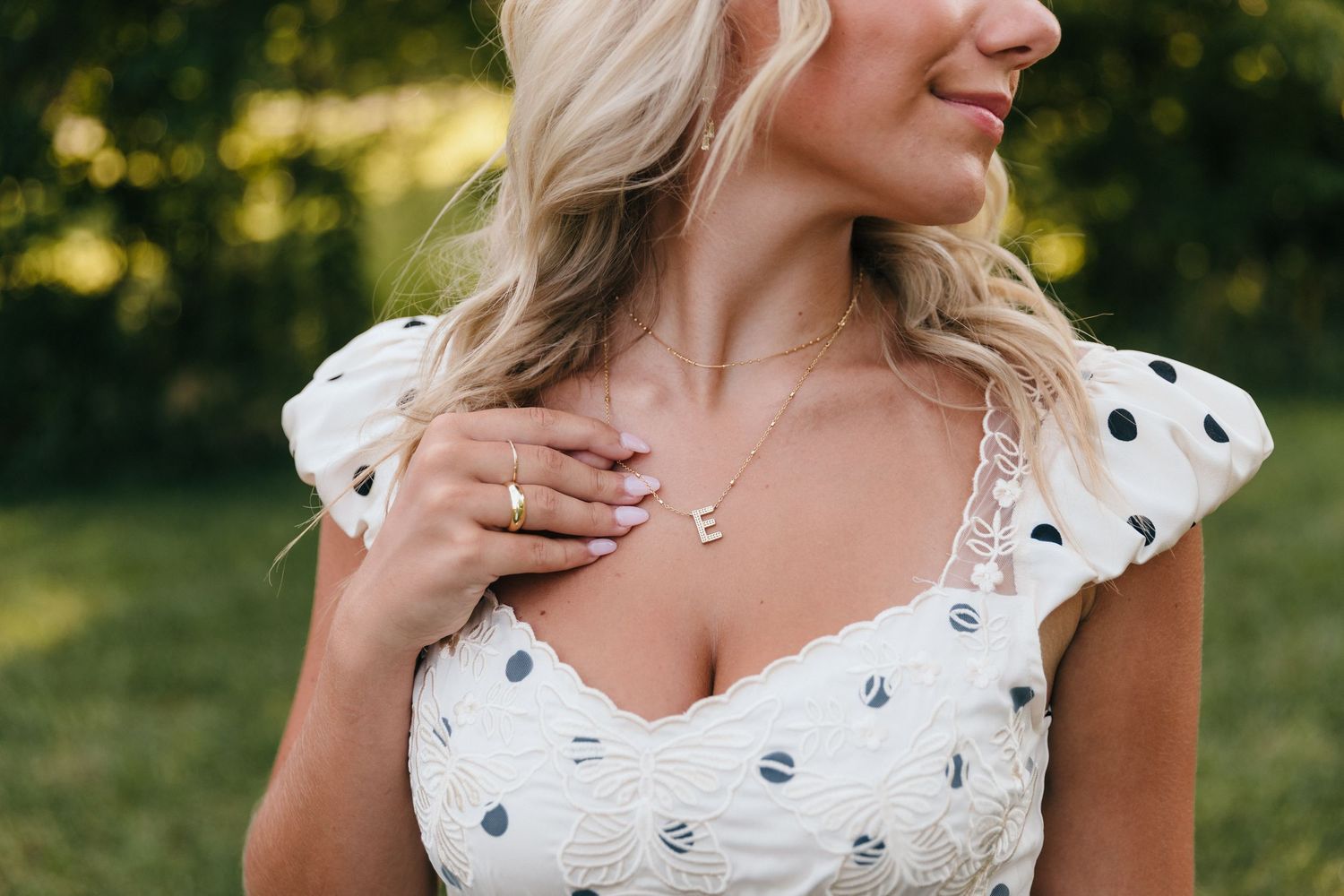 Blonde woman in white polka dot dress wearing gold letter E necklace and ring, posing outdoors.