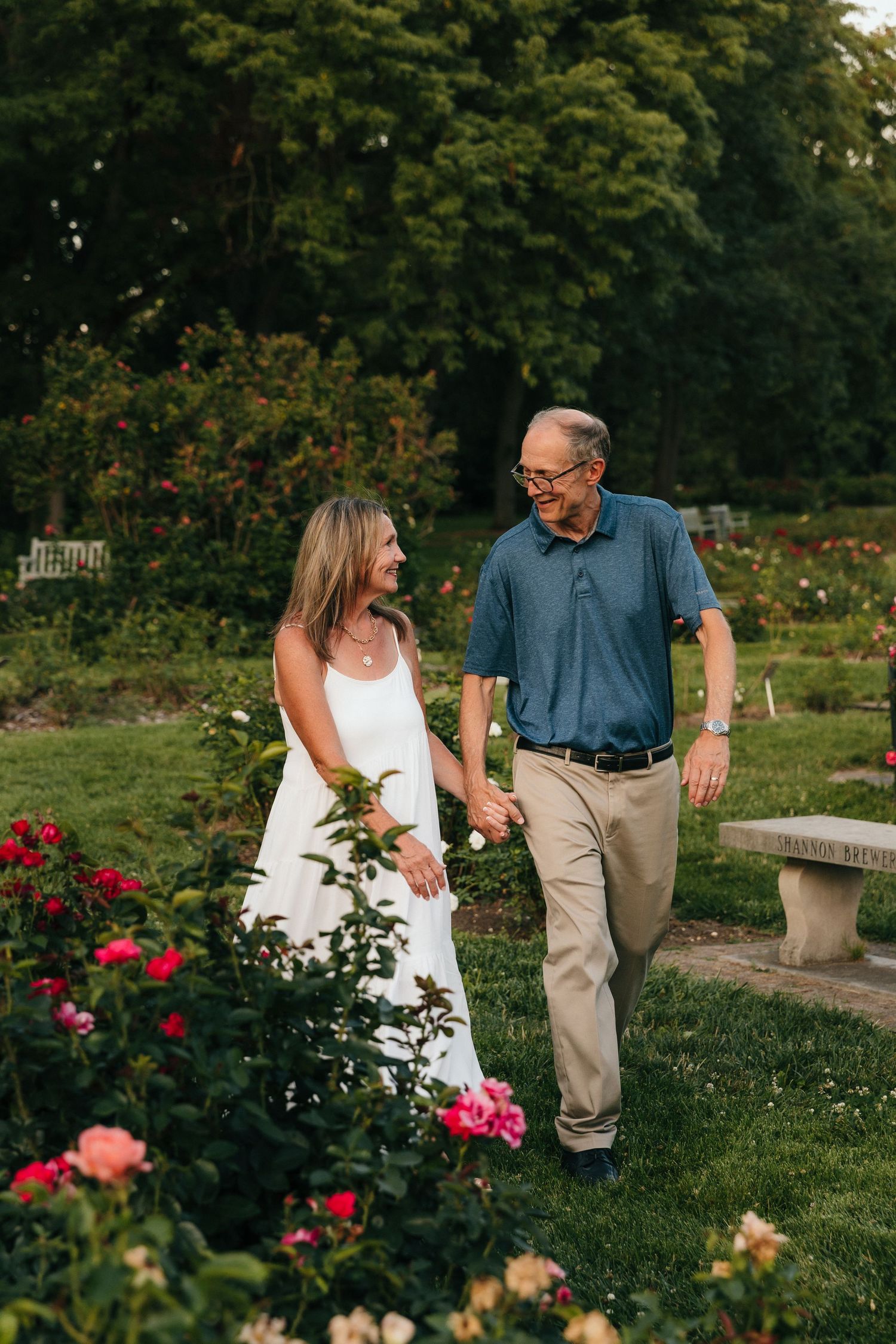 Couple holding hands and smiling while walking through a lush rose garden at dusk.