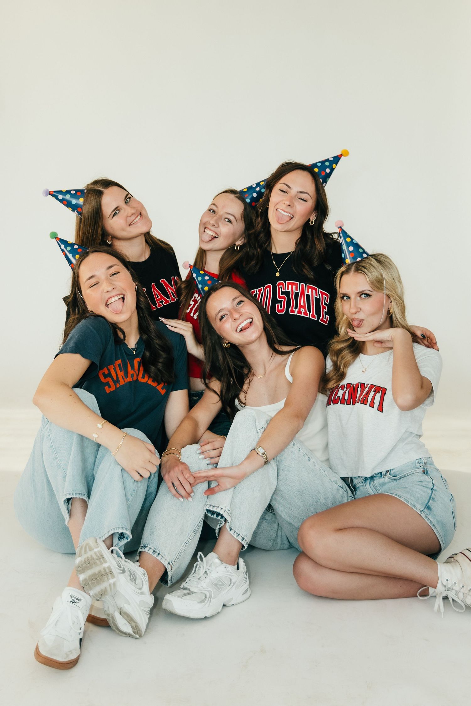 Six young women in college shirts and party hats pose together laughing on a white background.