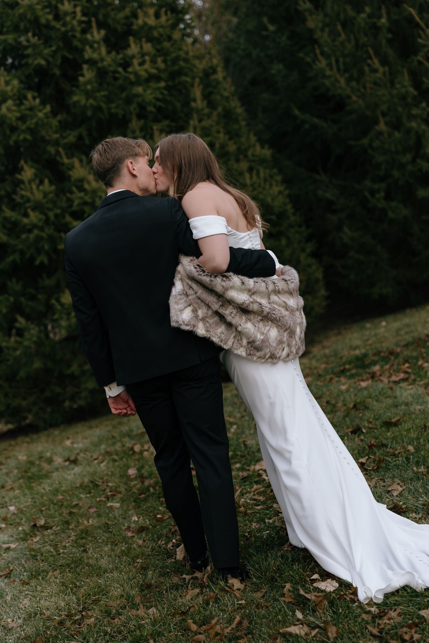 A romantic wedding couple embraces outdoors on a grassy hillside during an autumn day.