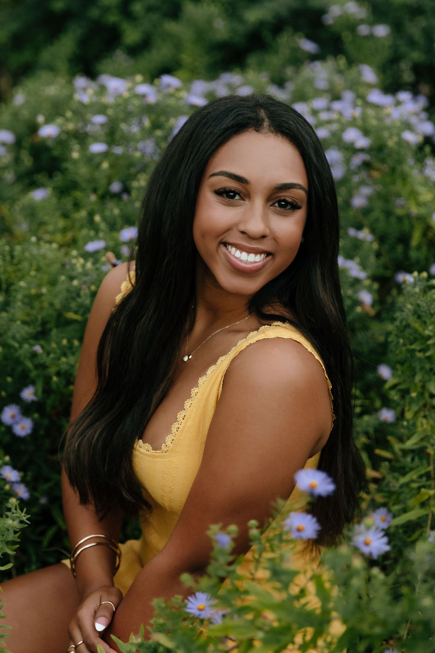 A woman in a yellow top poses outdoors among purple flowers in a garden setting.