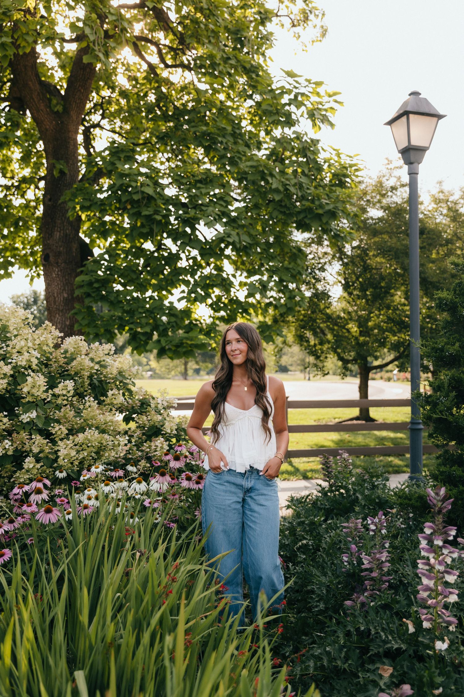 Young woman in white top and jeans posing among blooming flowers in a lush green park near a lamppost.