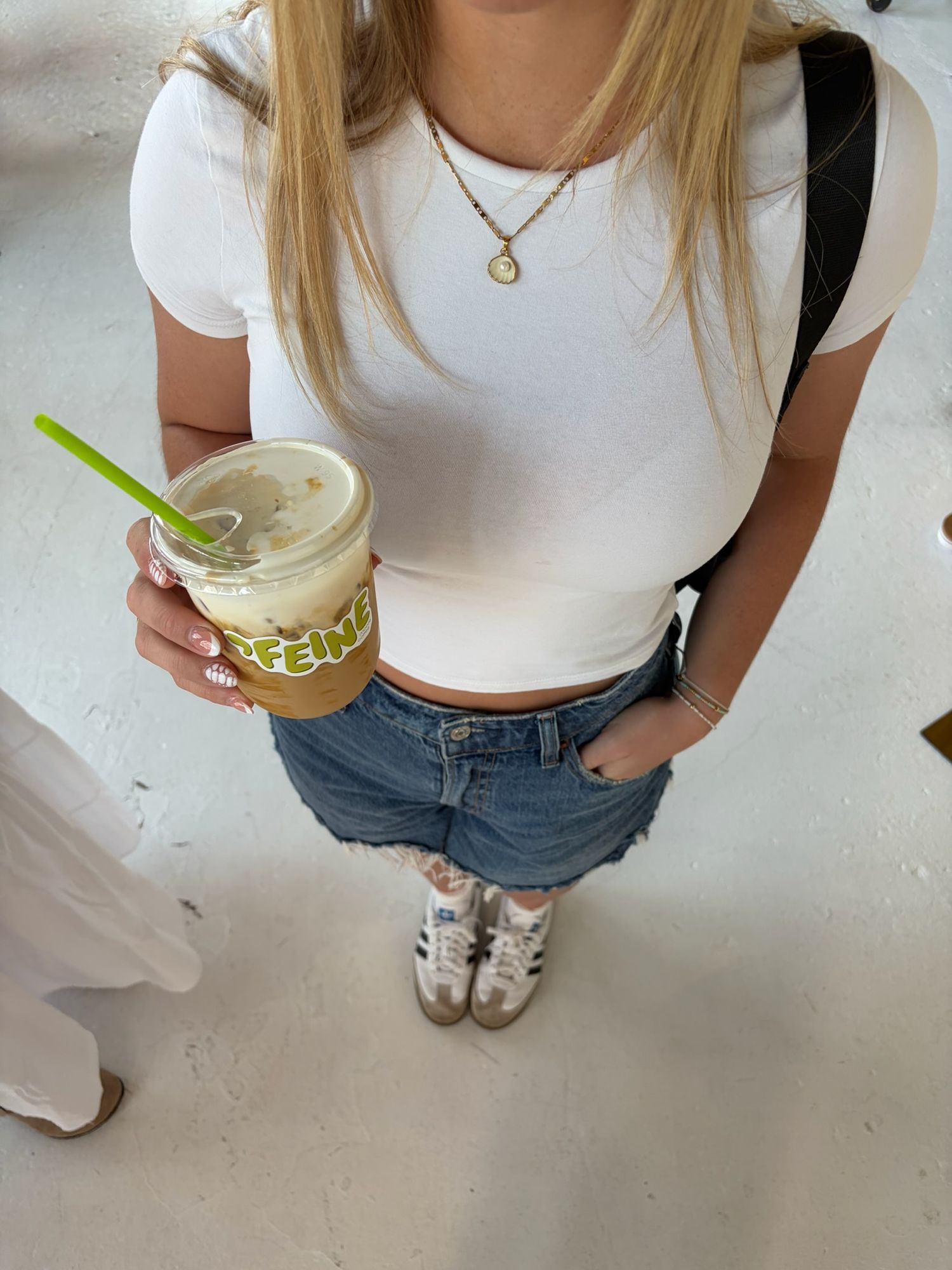 Young woman in white crop top and denim shorts holding an iced coffee drink with a green straw.