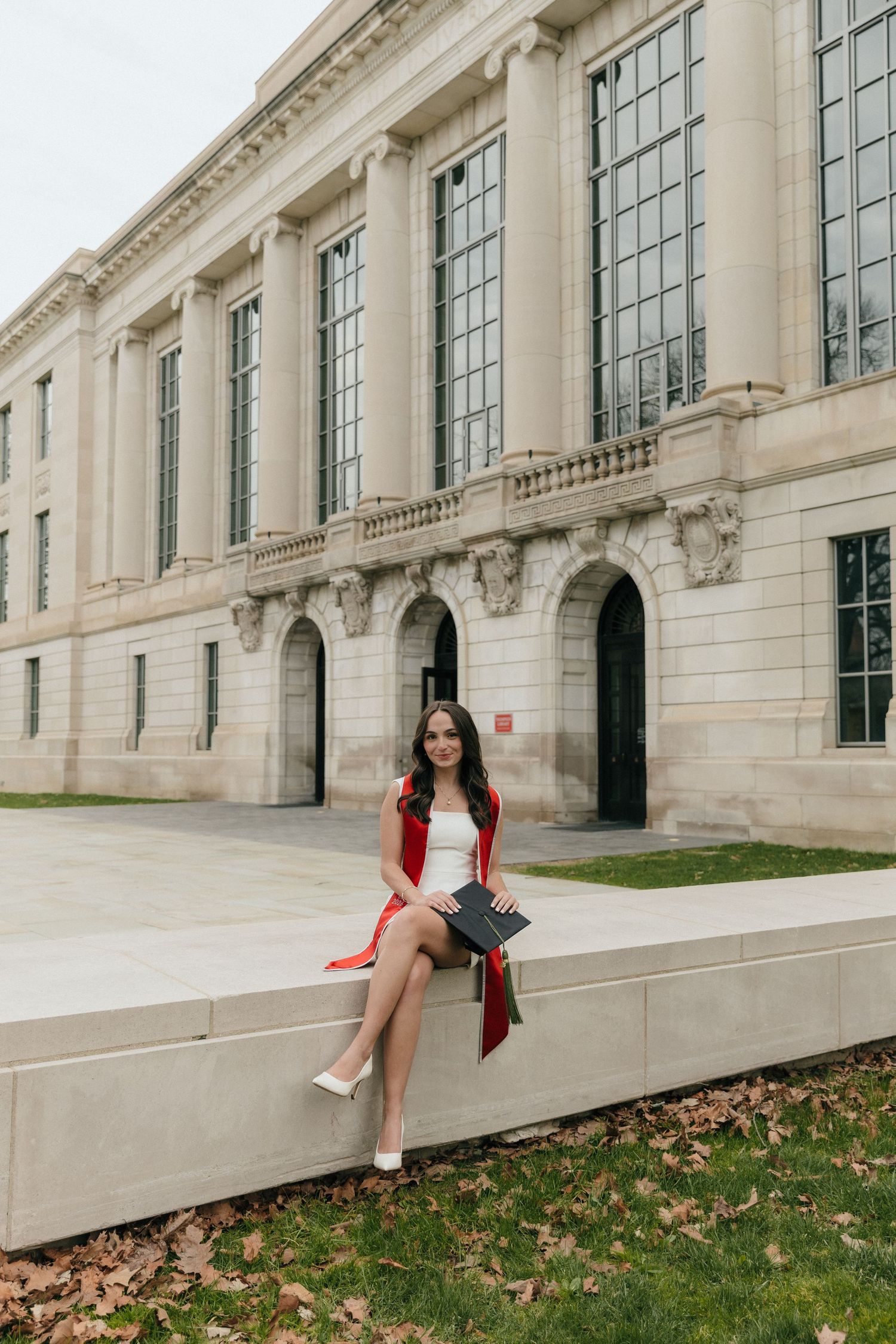 Female graduate in white dress and red stole sits on ledge holding cap outside neoclassical building.