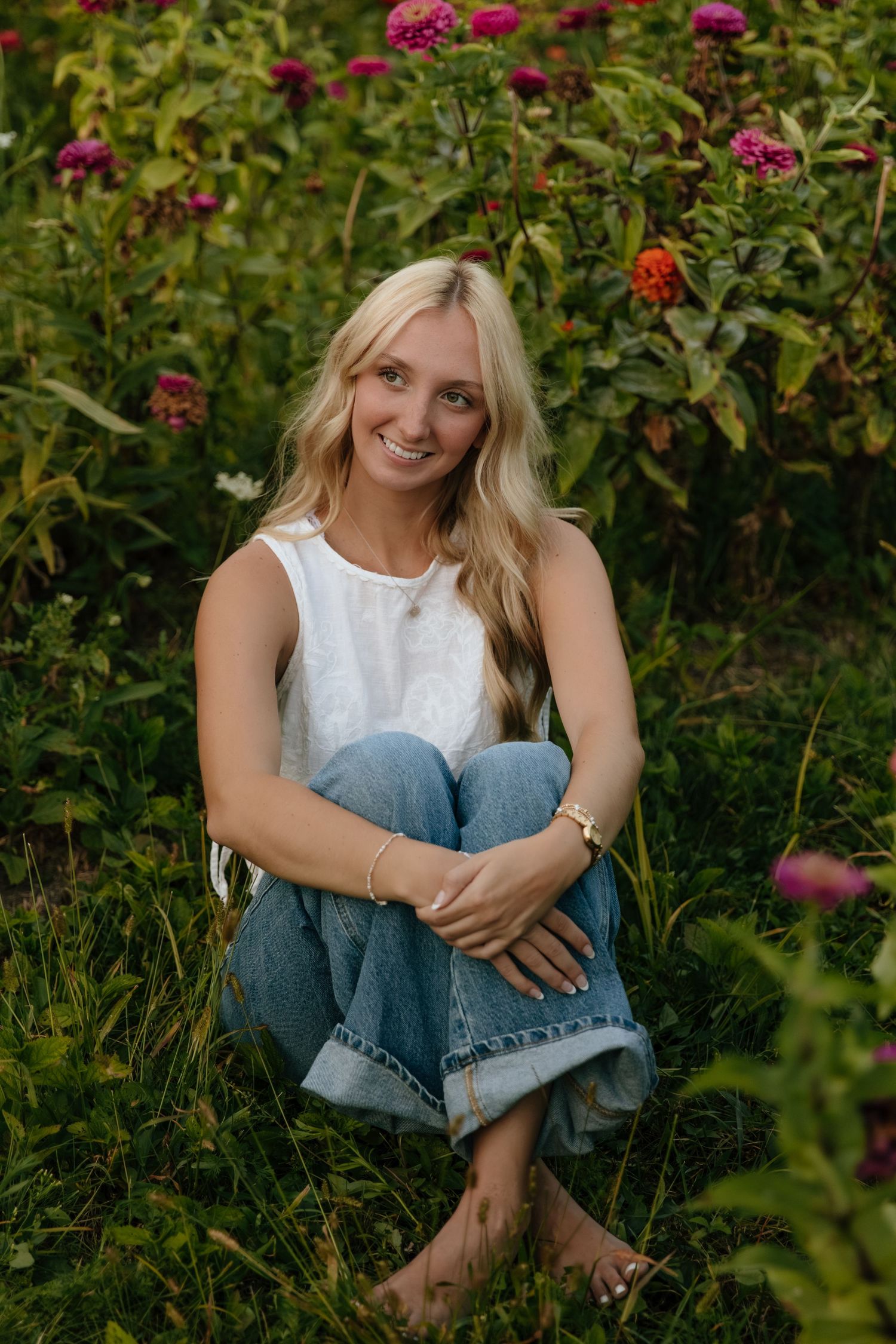 A woman in a white sleeveless top and jeans sits casually among blooming pink flowers in a garden setting.