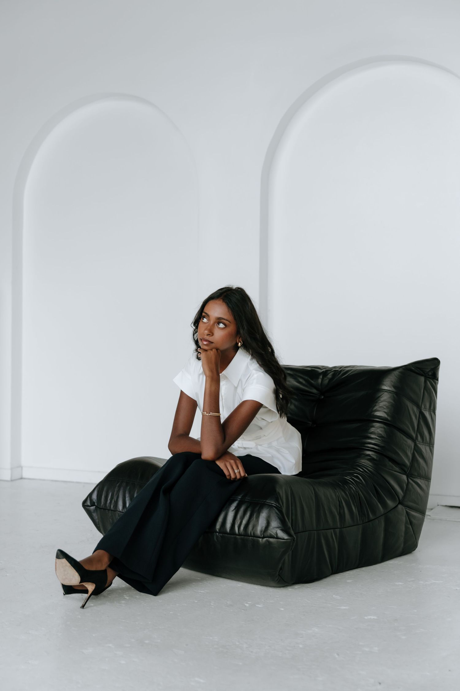 A person lounges elegantly on a black leather bean bag chair against a minimalist white backdrop in a fashion-forward pose.