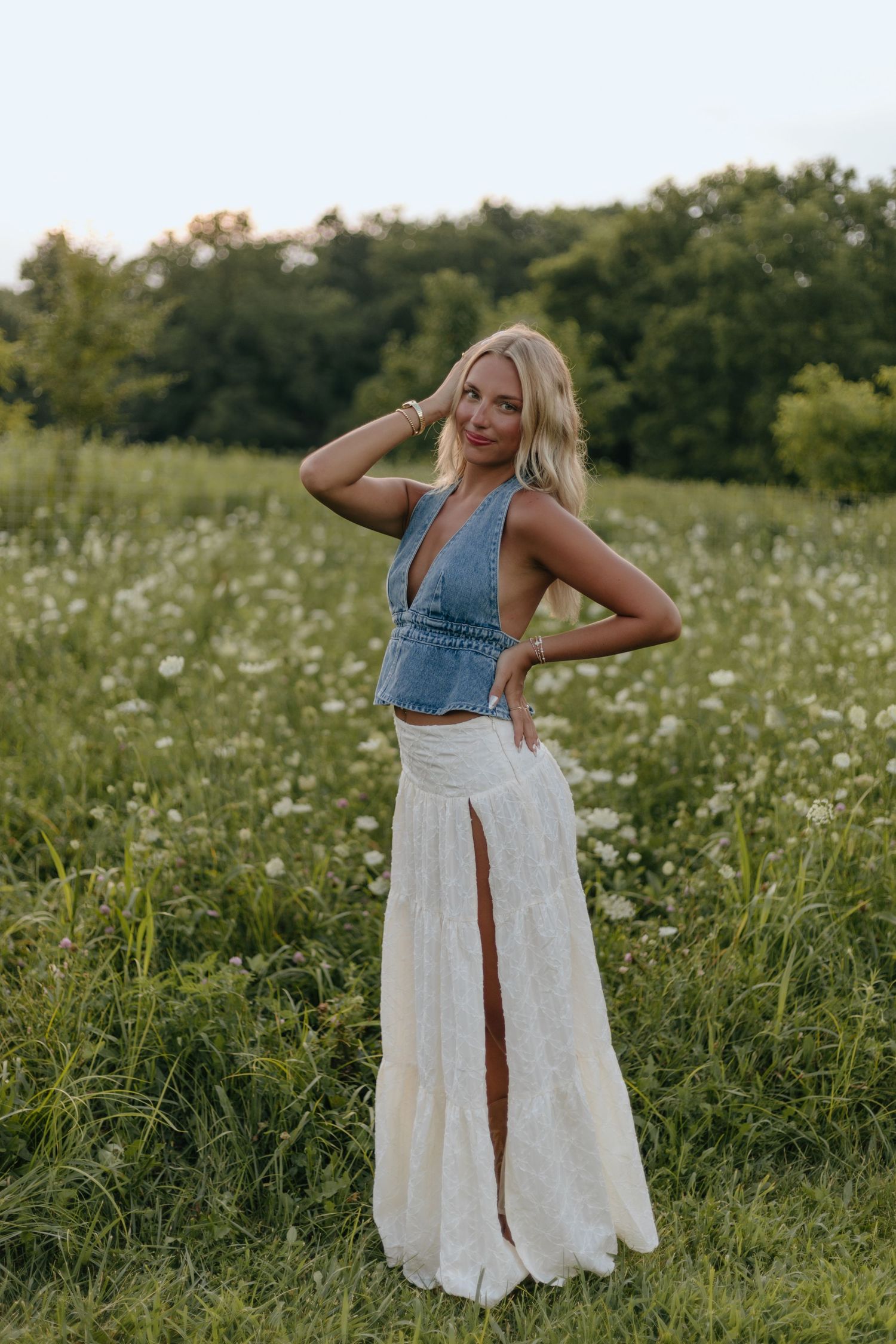 A person in a denim top and white maxi skirt poses playfully in a wildflower meadow at sunset.