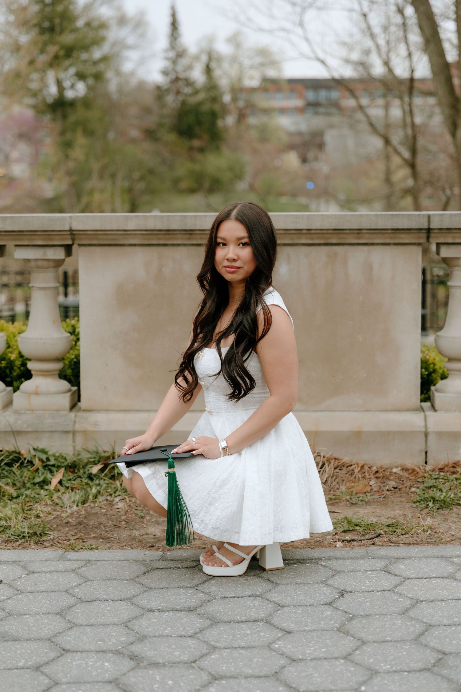 A graduate in a white dress sits on stone steps holding a graduation cap in an outdoor setting.