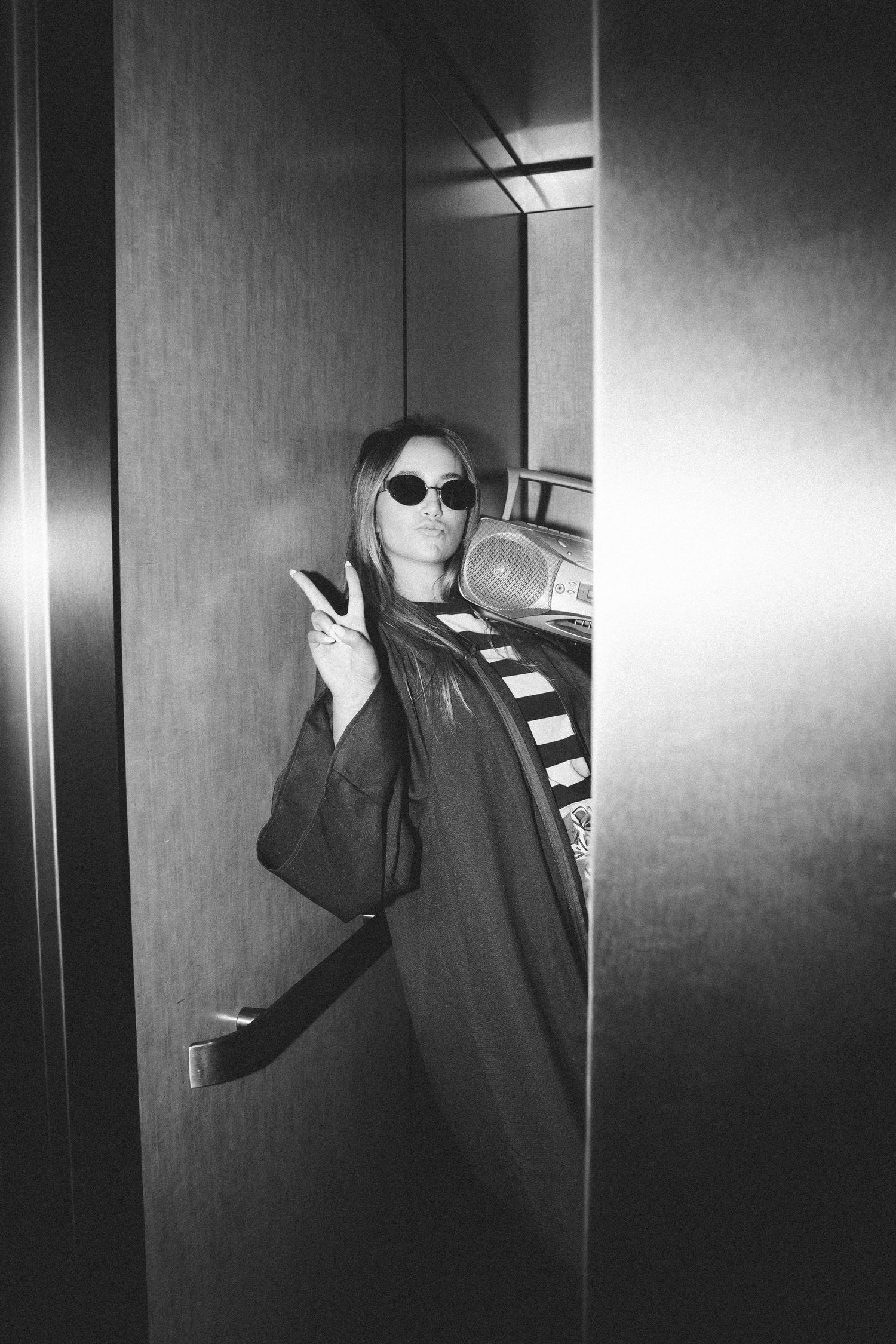 Black and white photo of a woman in sunglasses holding a boombox, flashing a peace sign in a doorway.