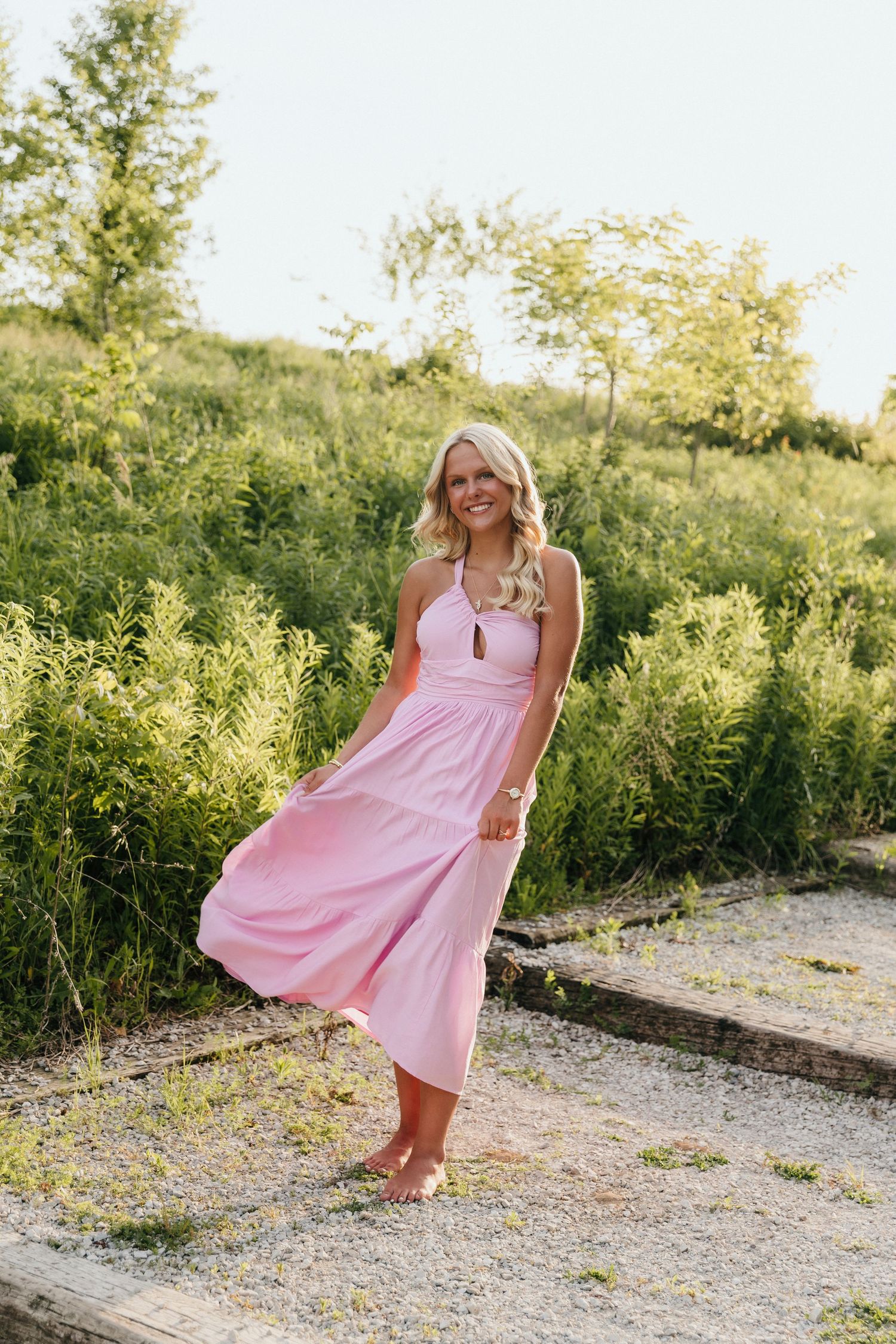 Smiling blonde woman in flowing pink midi dress standing barefoot outdoors surrounded by lush green foliage.