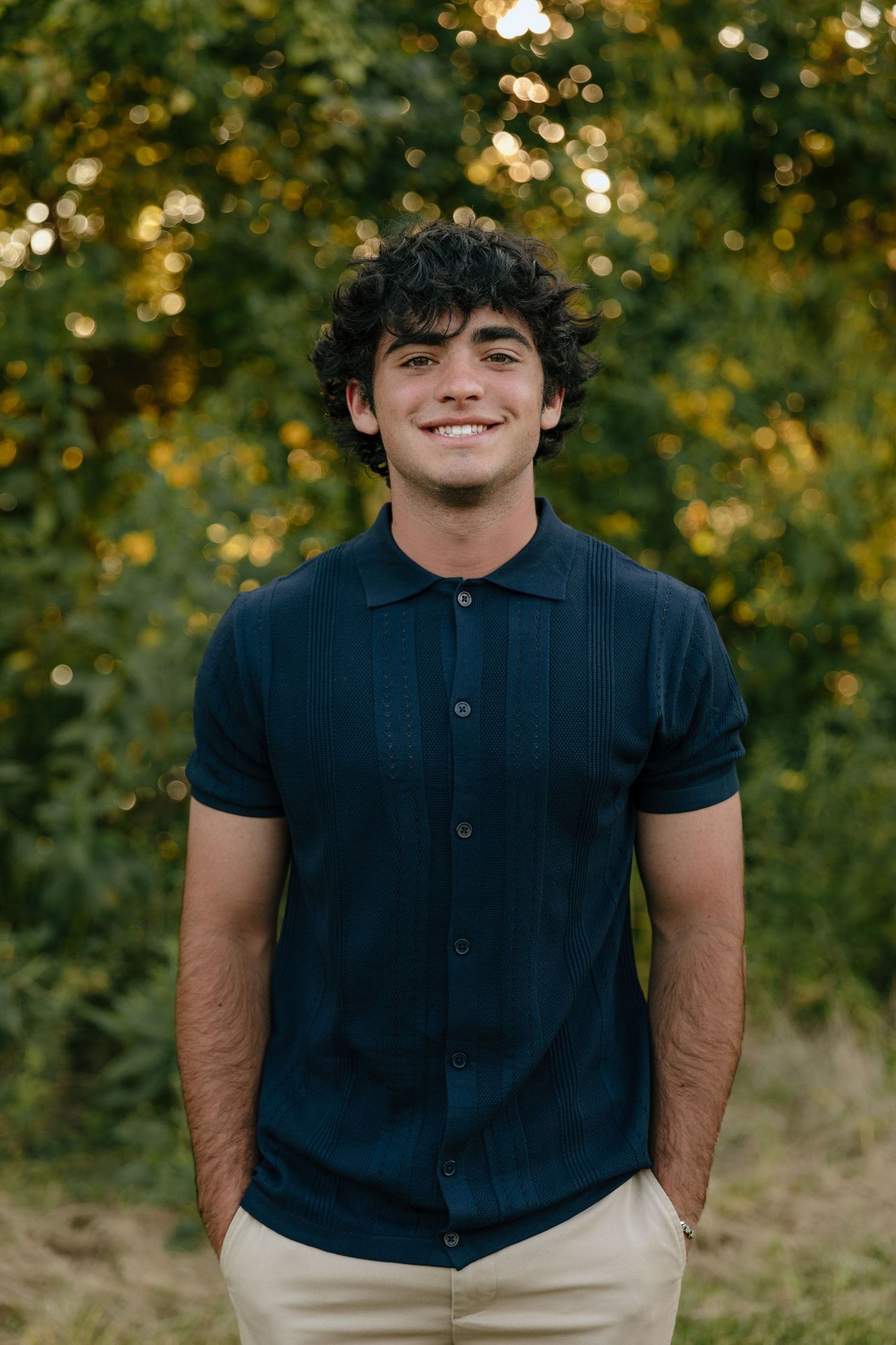 Portrait of someone wearing a navy blue polo shirt against a blurred natural background with golden bokeh lights.