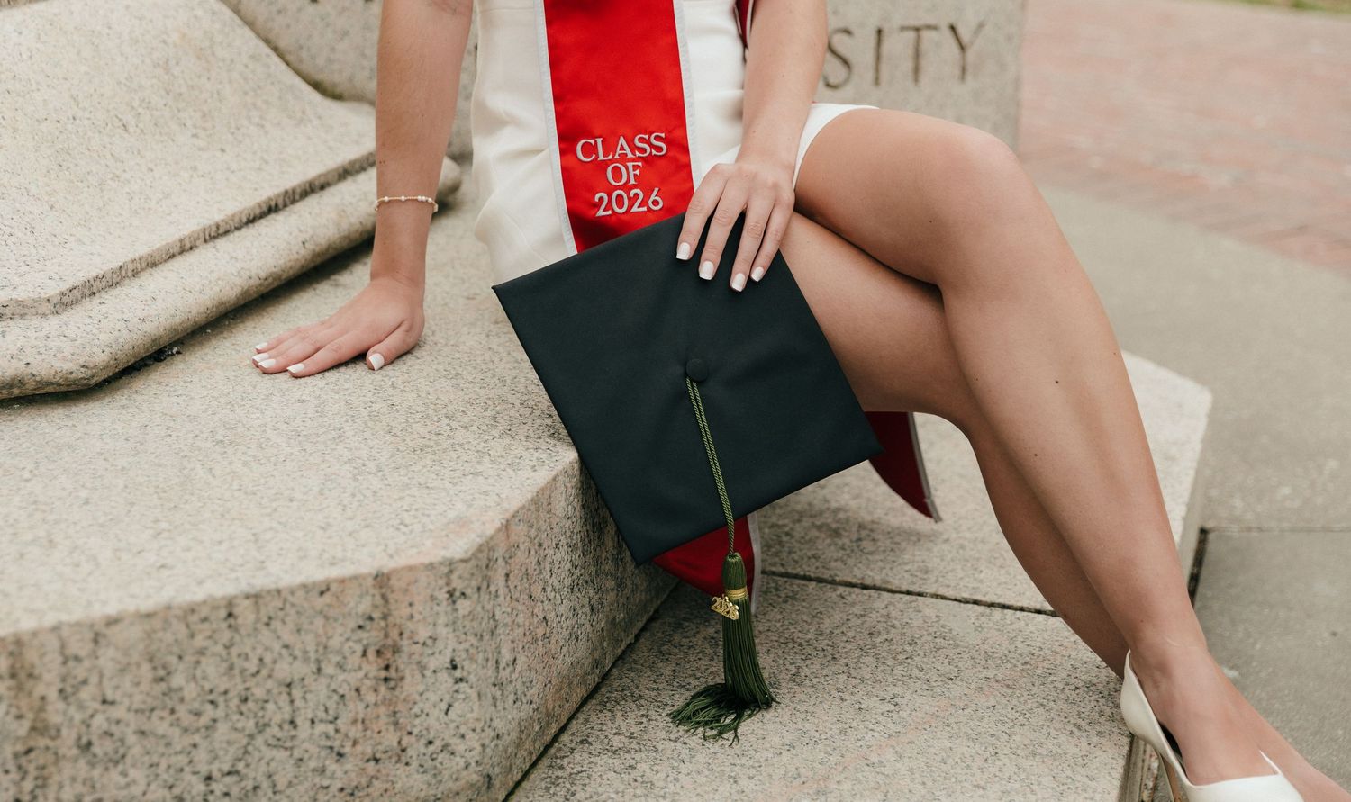 College graduate sits on stone steps holding a Class of 2026 cap with red tassel and graduation sash.