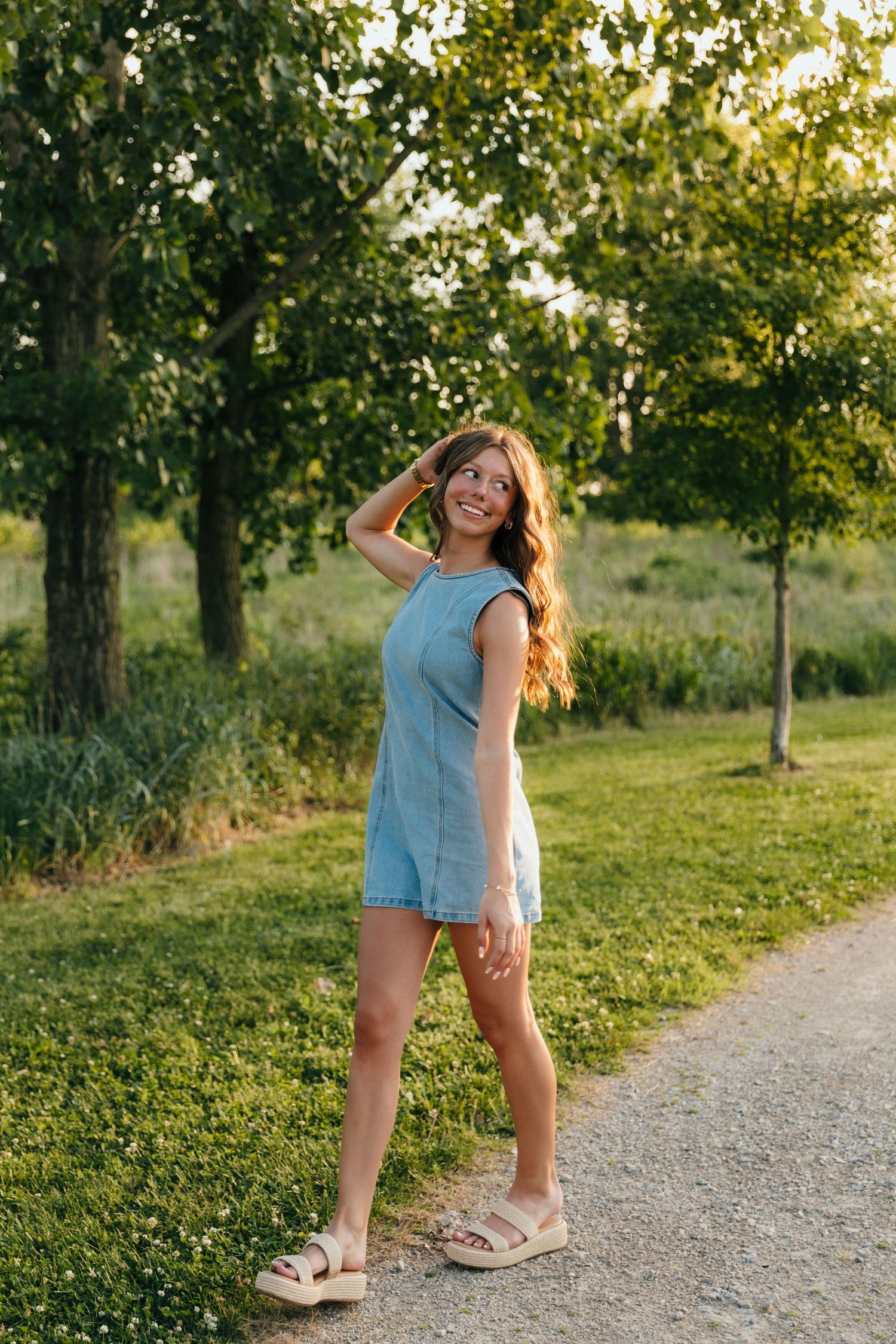 Young woman in denim romper smiling outdoors on a sunny day, surrounded by lush green trees and grass.