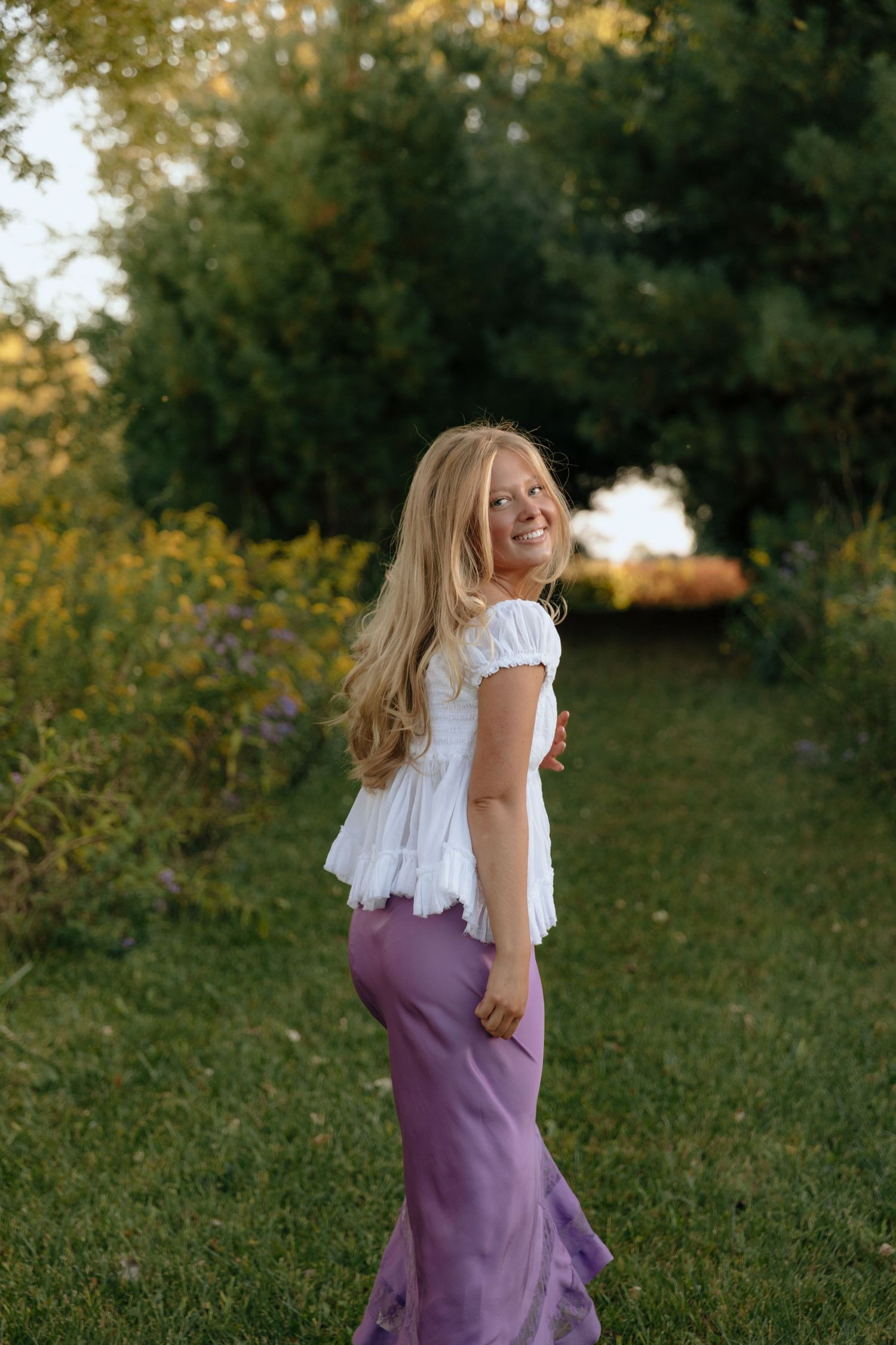 A person in a purple dress spins joyfully in a grassy outdoor setting during golden hour.