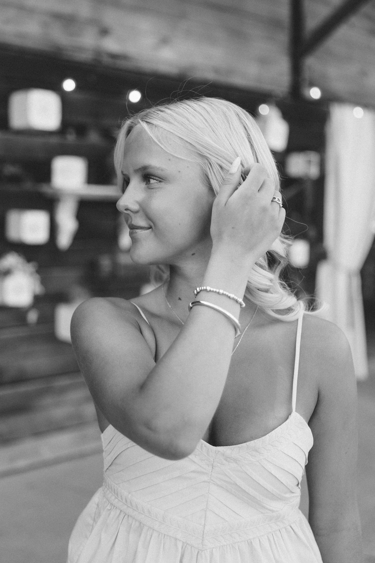 Black and white photo of a smiling blonde woman in a white sundress, touching her hair, wearing bracelets indoors.
