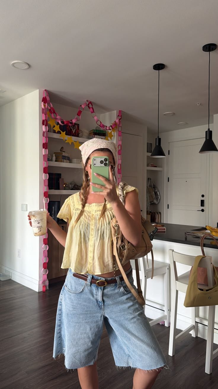 Woman in yellow top, denim shorts, brown belt and bag, taking mirror selfie in modern kitchen.
