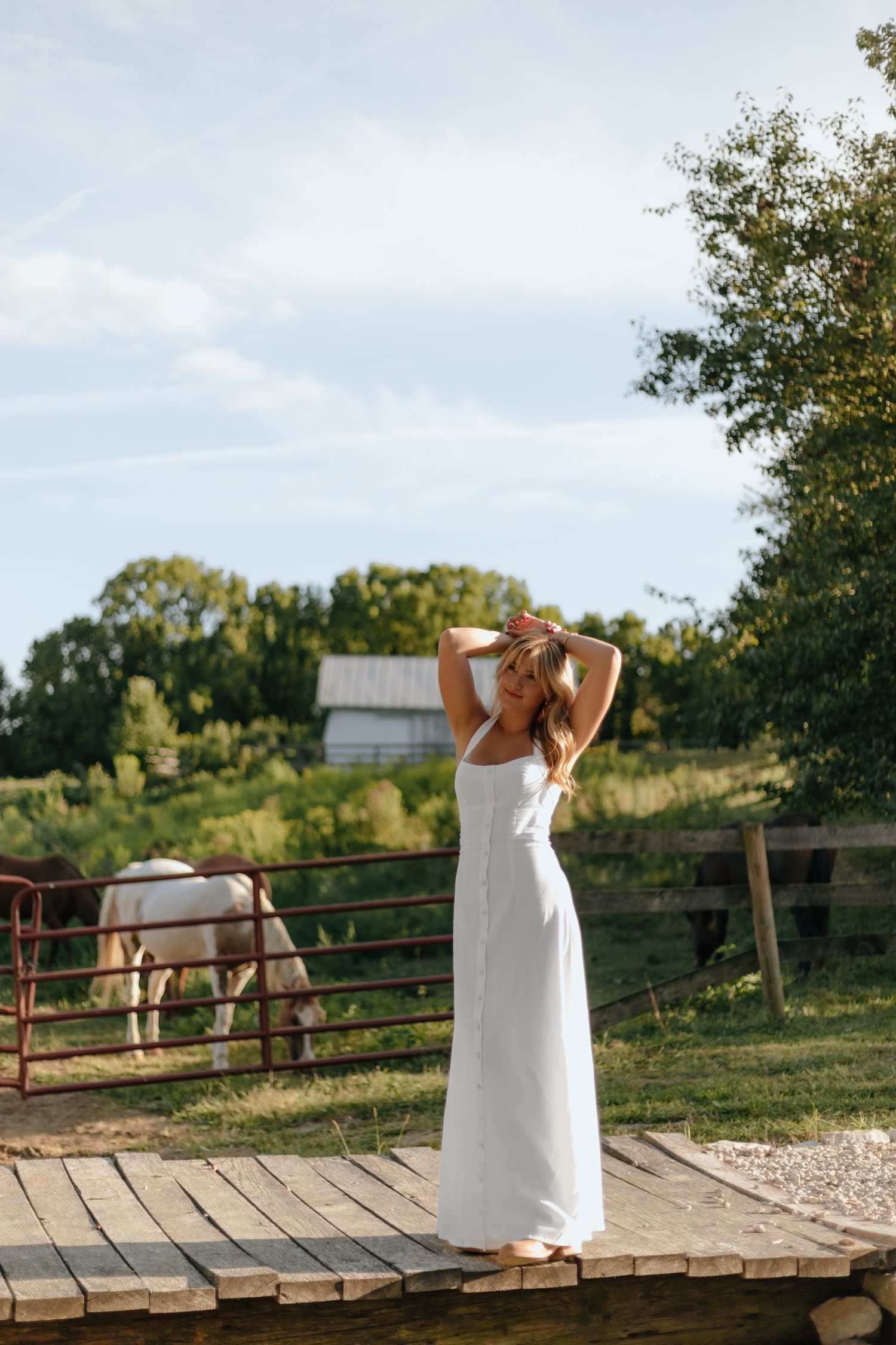 Woman in white dress stands on wooden deck with farm and horse in background.
