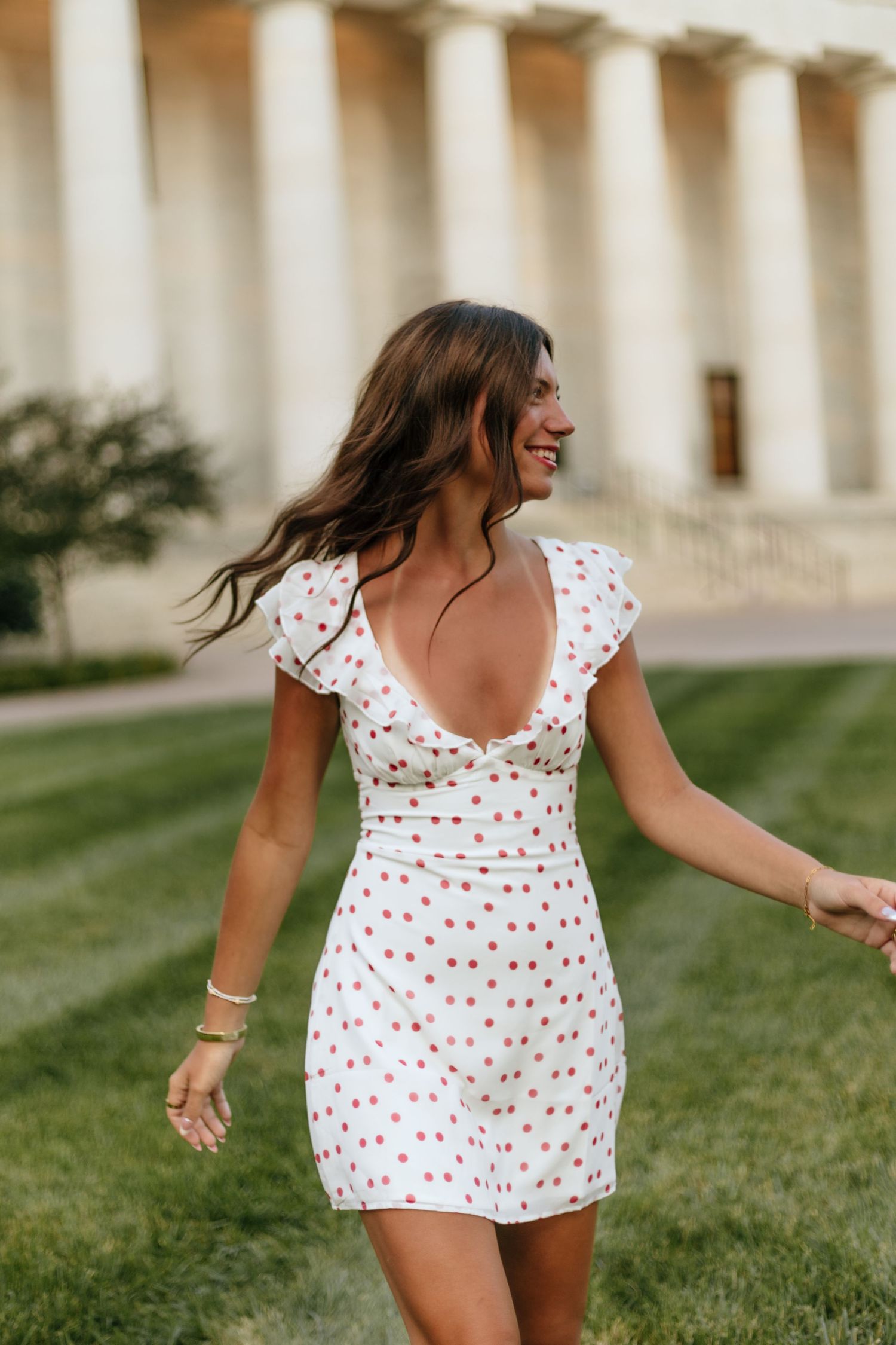 Person in white polka dot dress twirling playfully on grass near classical architecture.