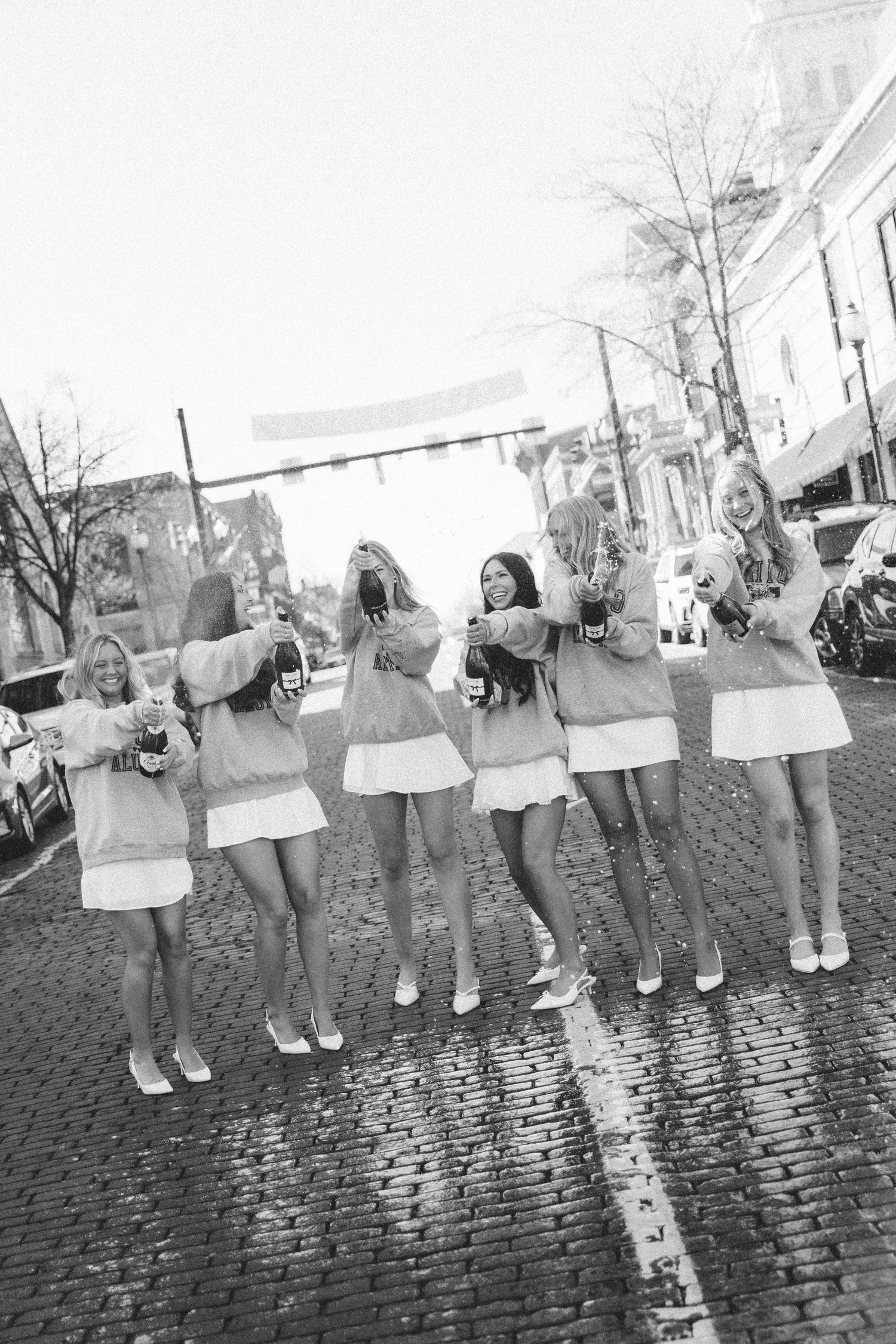 Six young women in matching outfits celebrate on a cobblestone street, holding champagne bottles in a black and white photo.