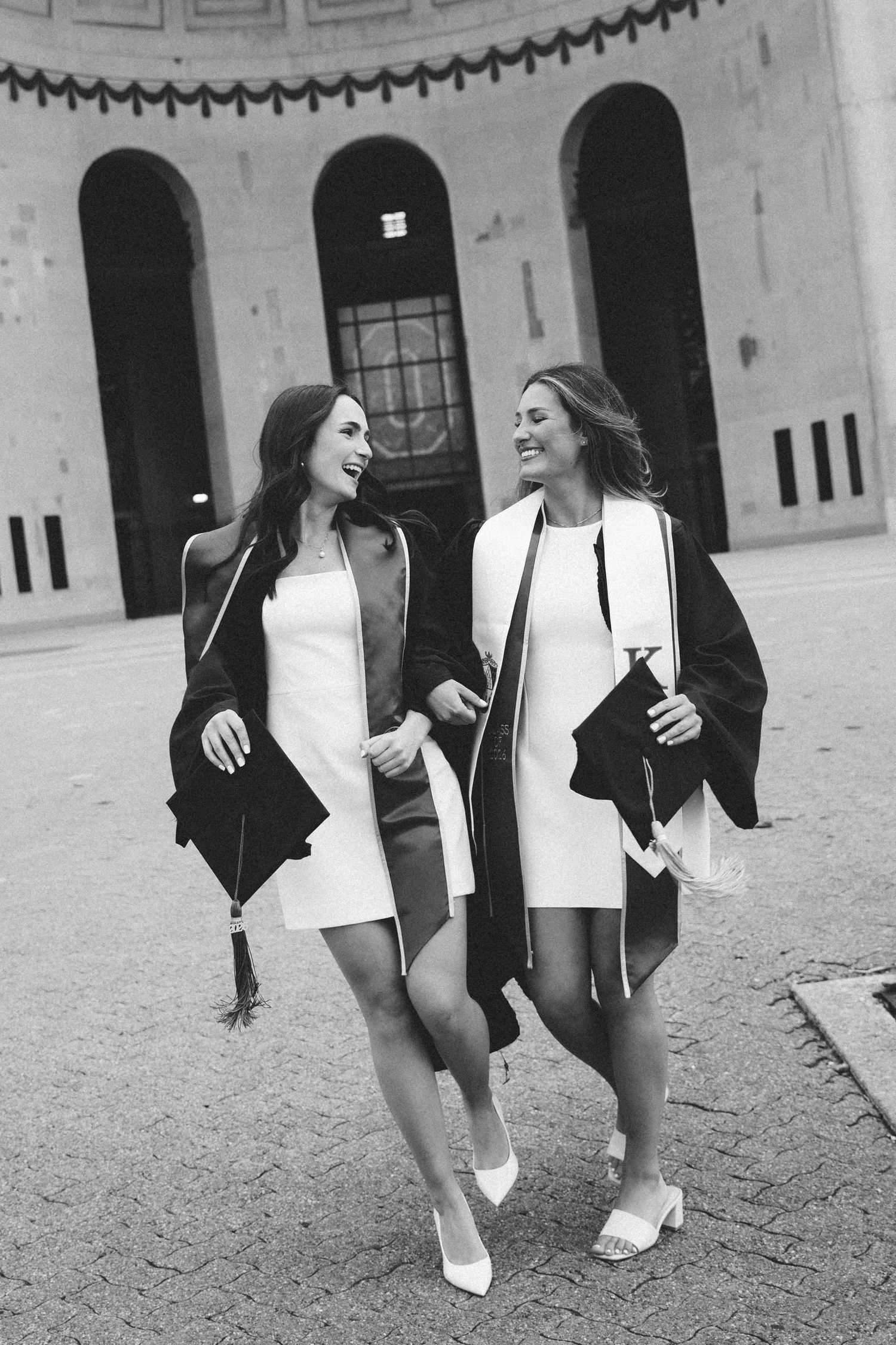 Two smiling female graduates in white dresses and black robes walking together in black and white photo.
