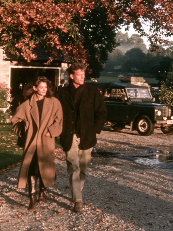 A man and woman in coats walk along a gravel path past a vintage green Land Rover and a stone cottage amid autumn foliage.