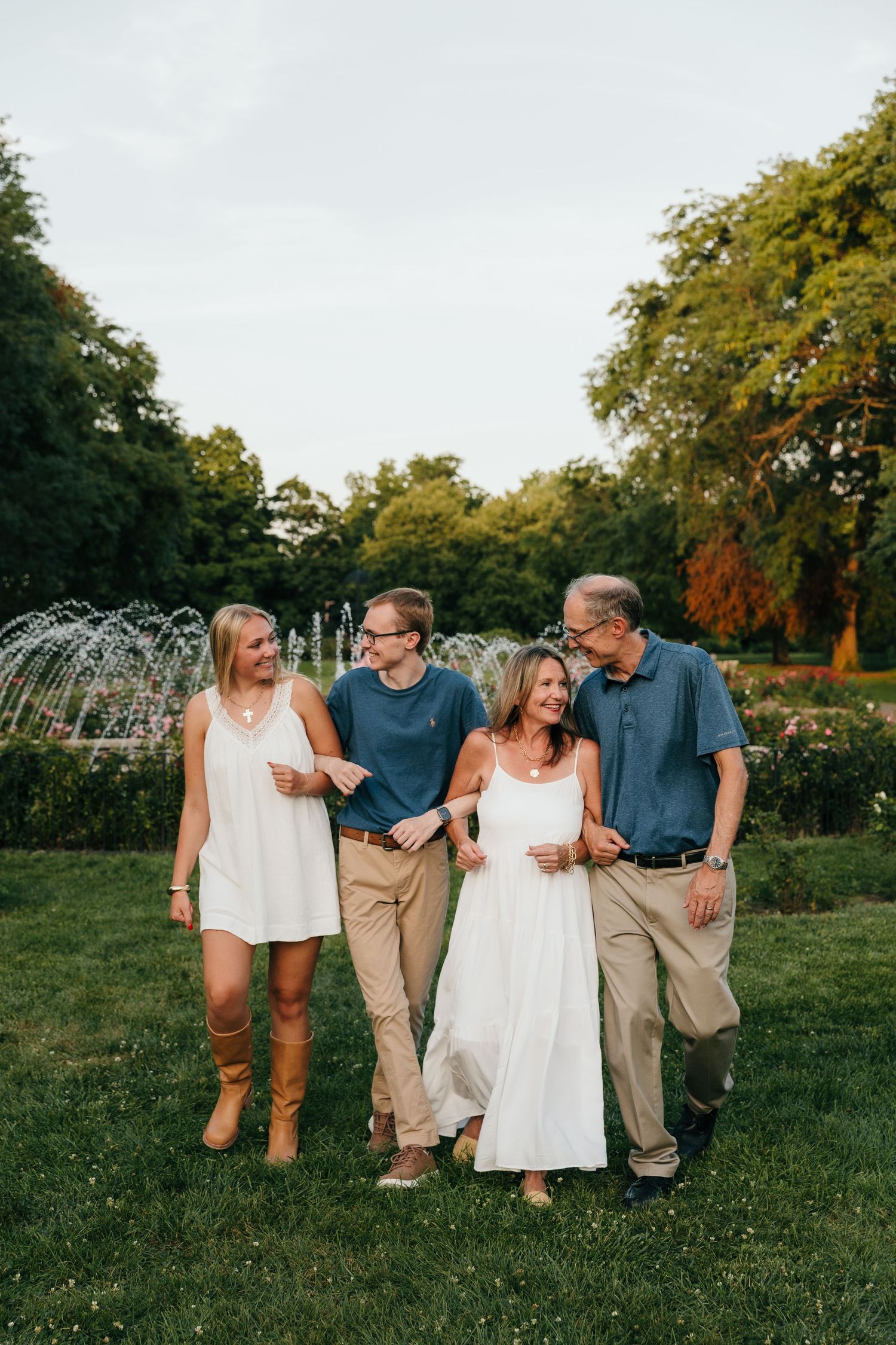 Family of four smiling and walking together in a lush green park with fountains in the background.