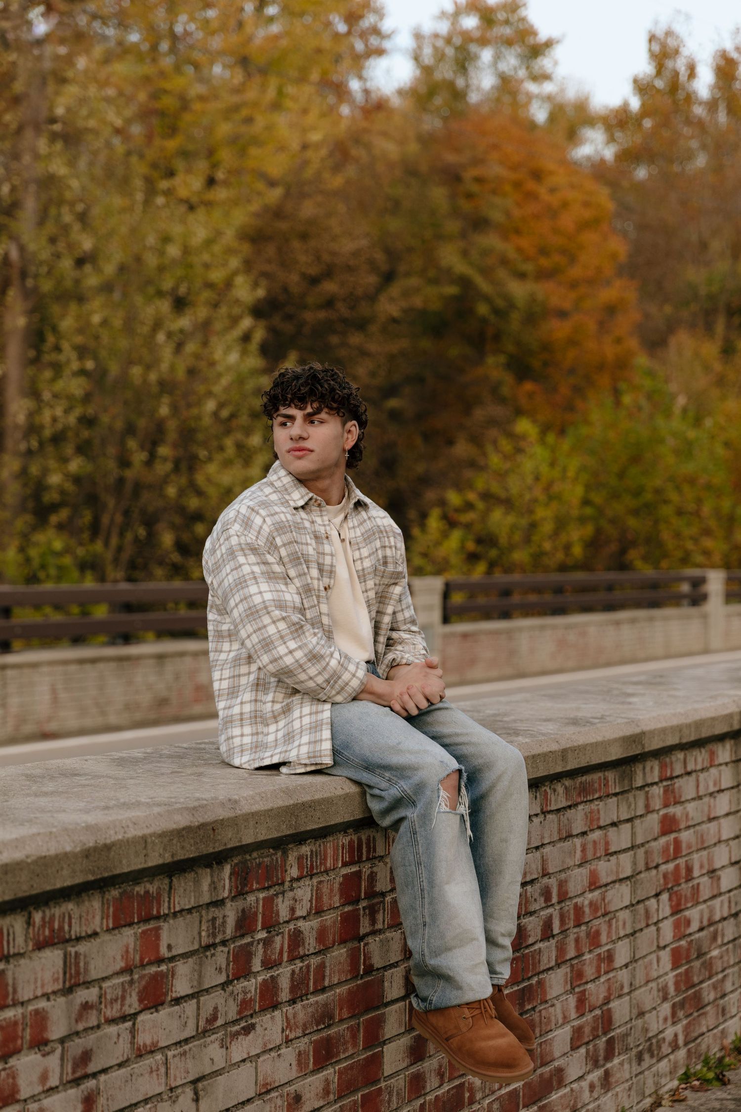 Person in plaid shirt and jeans sits on brick wall surrounded by autumn foliage during outdoor photoshoot.
