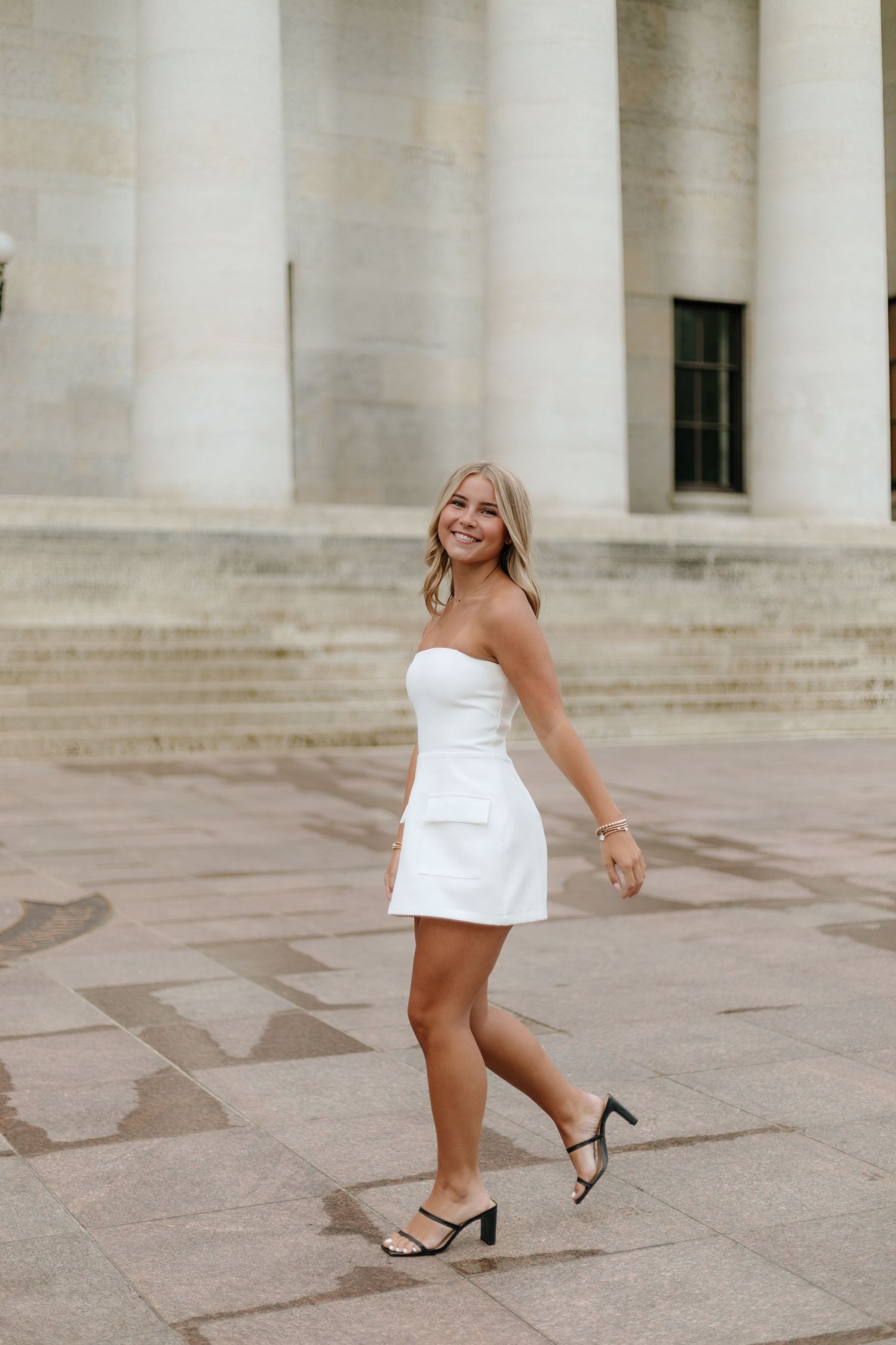 Someone in a white strapless mini dress and heels poses playfully on stone steps near classical columns.