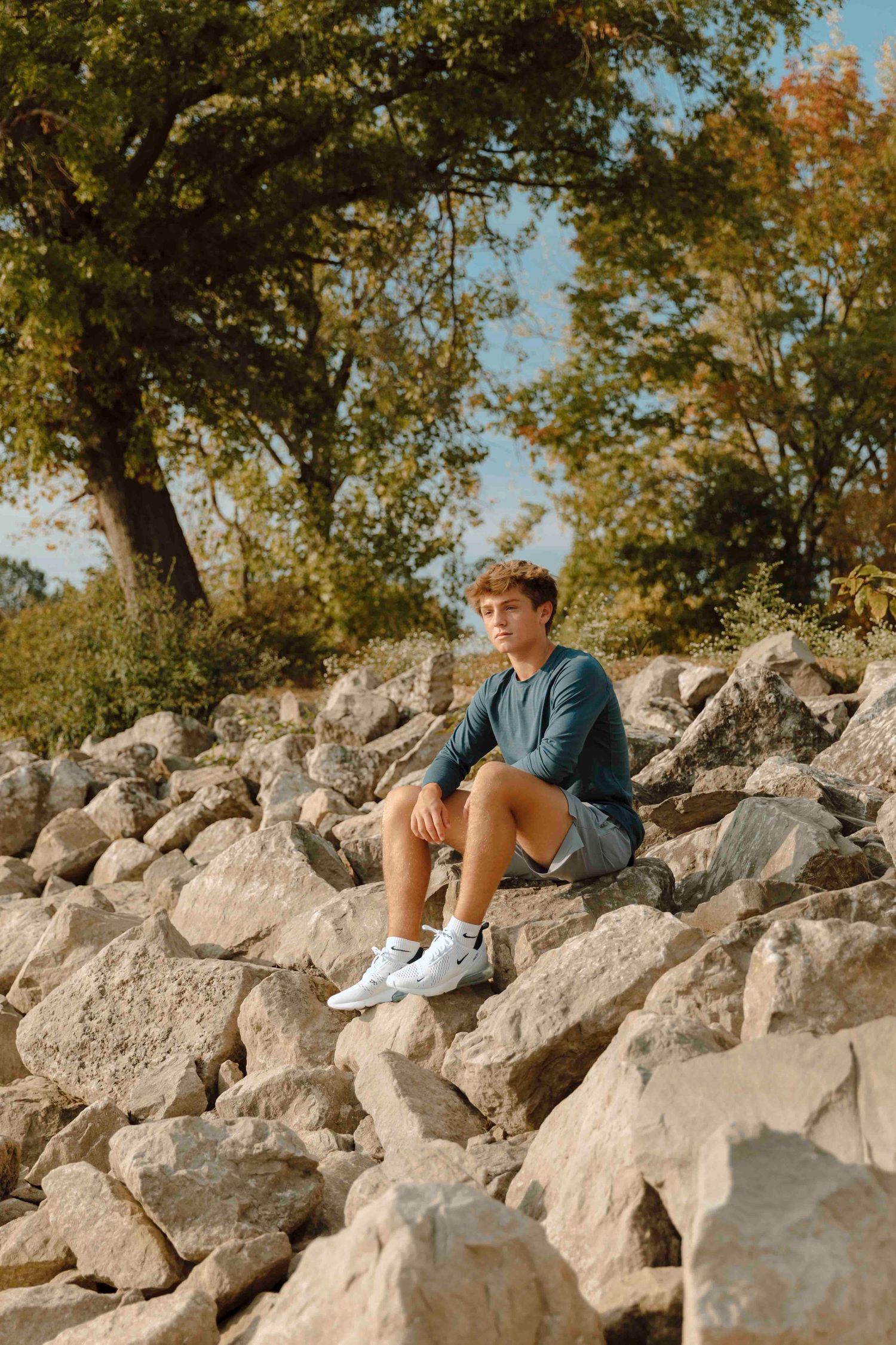 Young man in blue shirt and gray shorts sitting on large rocks outdoors with trees in background.