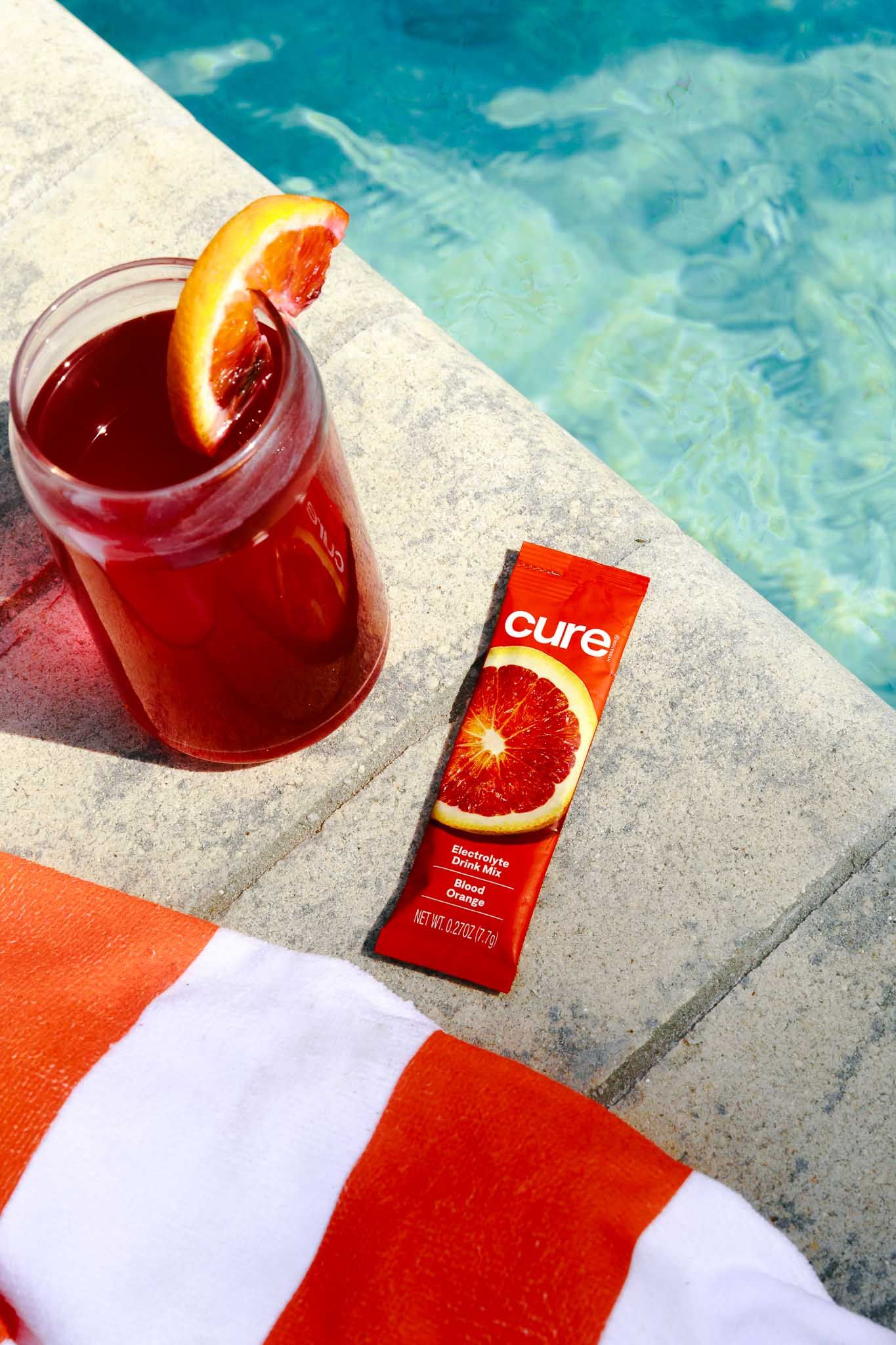 Red cocktail with orange garnish next to Cure drink mix packet by a swimming pool.