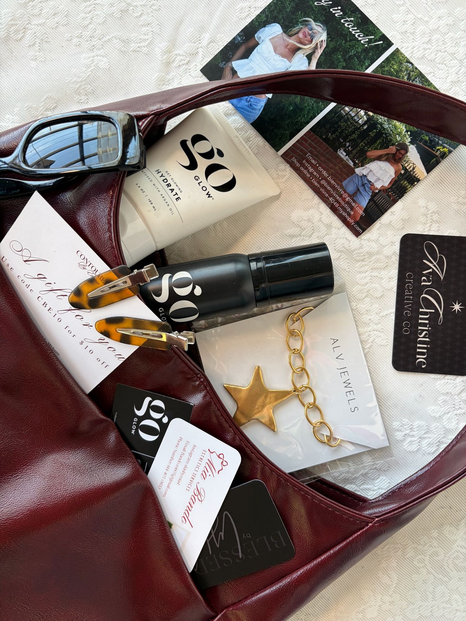 Burgundy leather handbag filled with beauty products, gold jewelry, hair clips, sunglasses, and promotional cards on white surface.