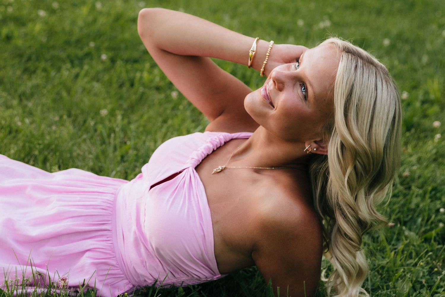 Blonde woman in pink dress lying on green grass, wearing gold jewelry, looking up with a serene smile.