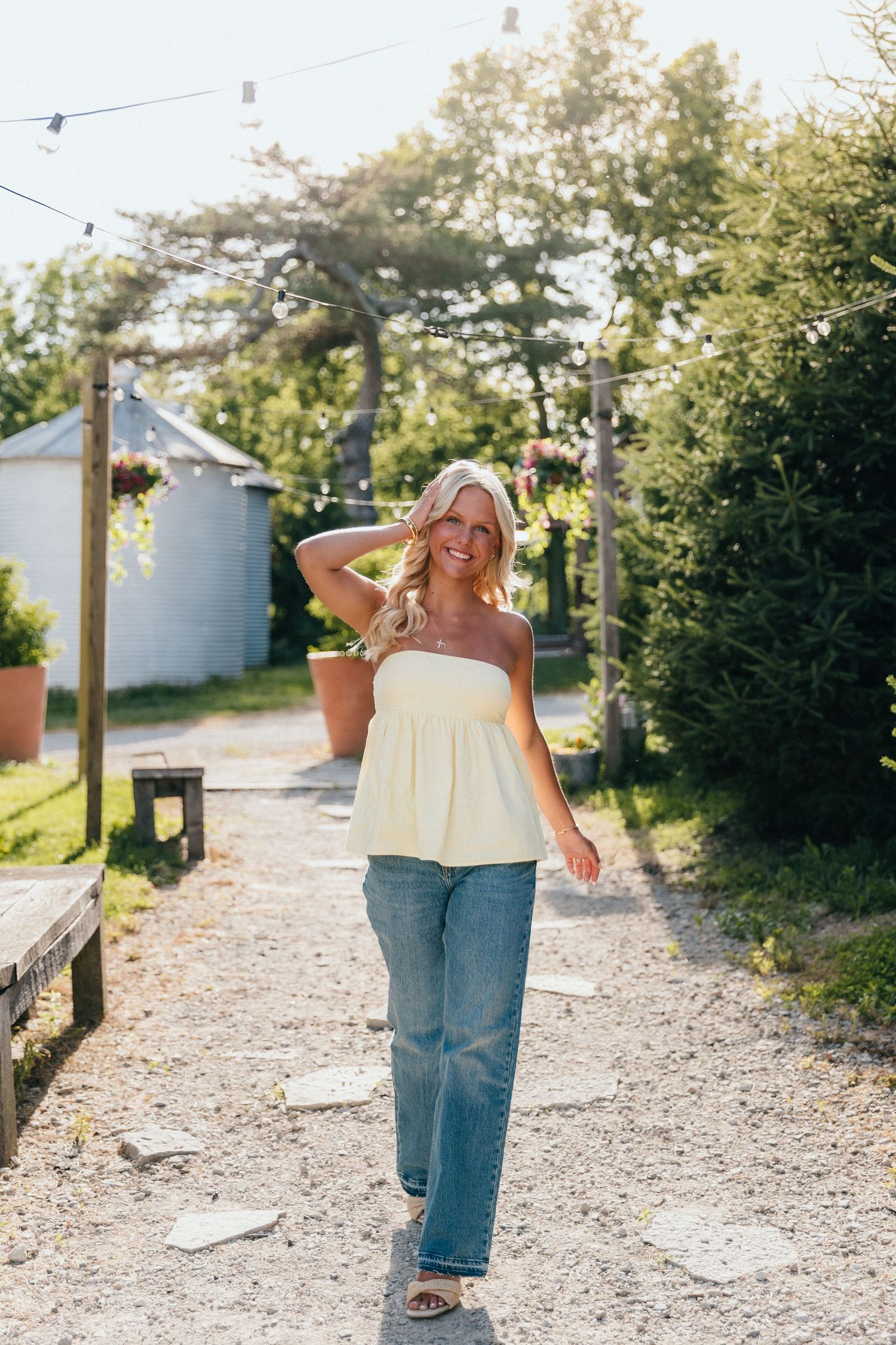 Woman in yellow strapless top and wide-leg jeans walking on a gravel path surrounded by lush greenery.