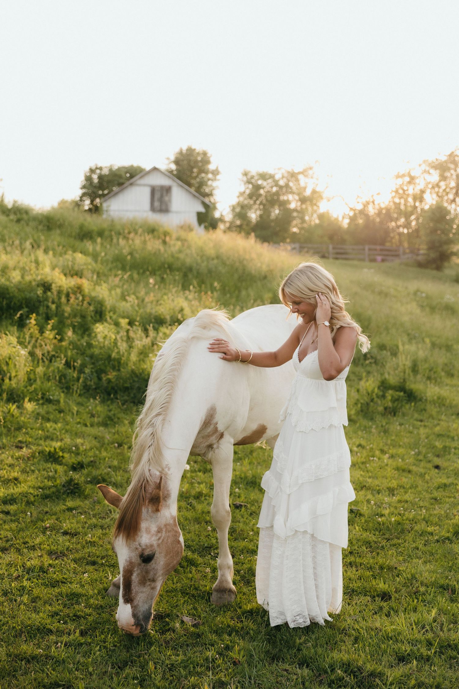 Blonde woman in white ruffled dress petting a white horse grazing in a sunlit green field with a farmhouse backdrop.