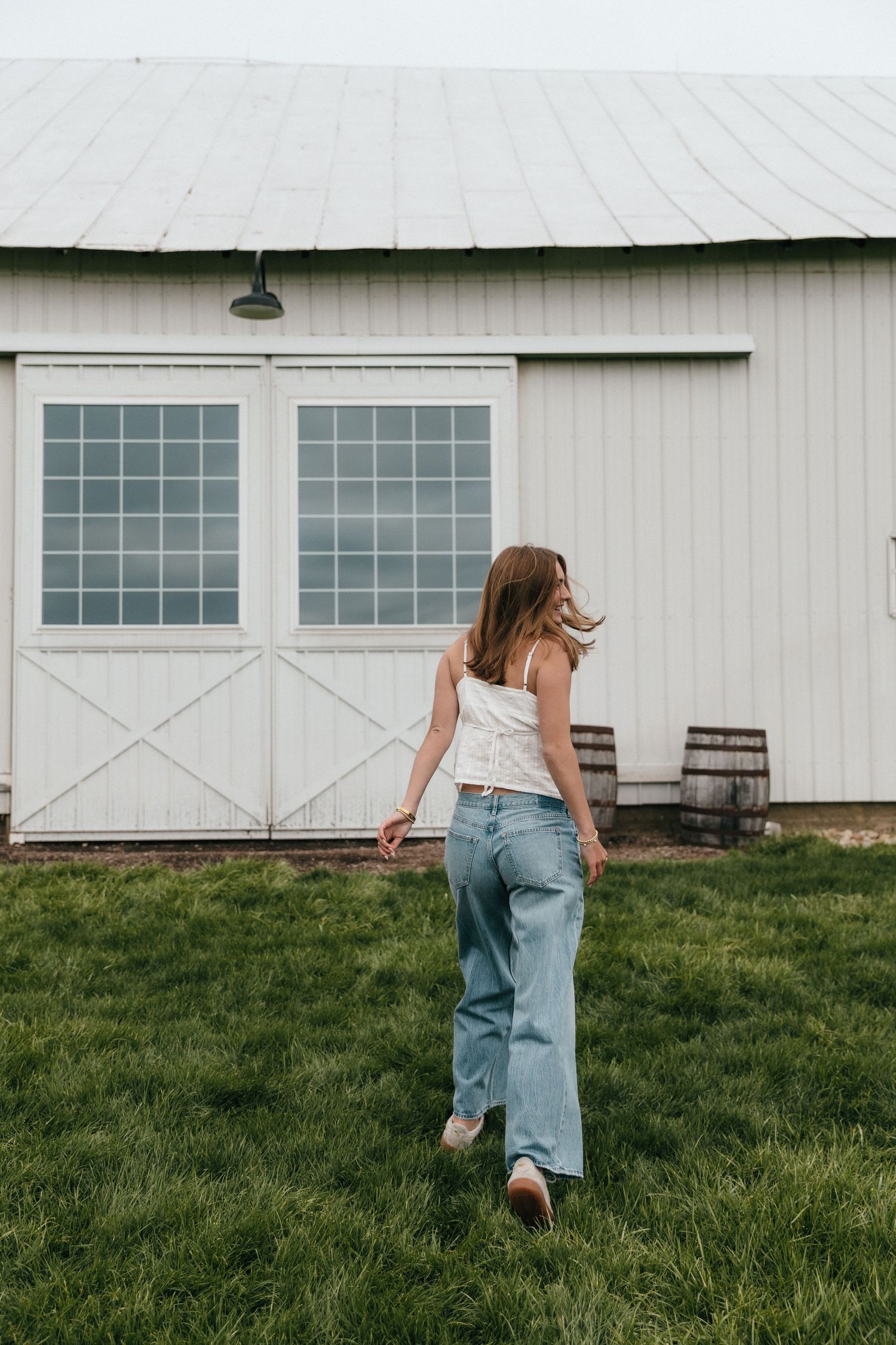 Young woman in white top and wide-leg jeans walking toward a white barn on a green lawn.