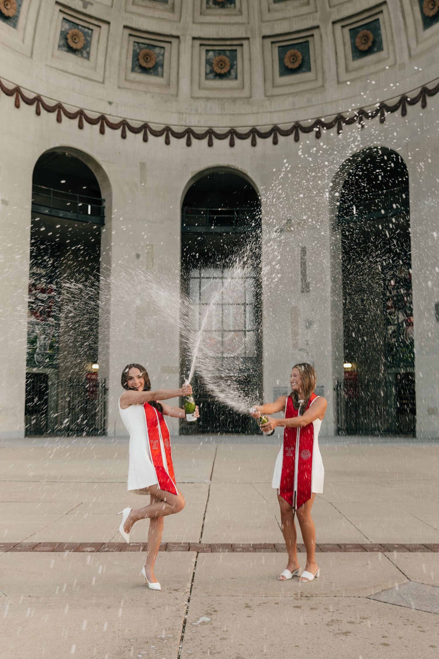 Two female graduates spray champagne bottles in celebration inside a grand domed building.