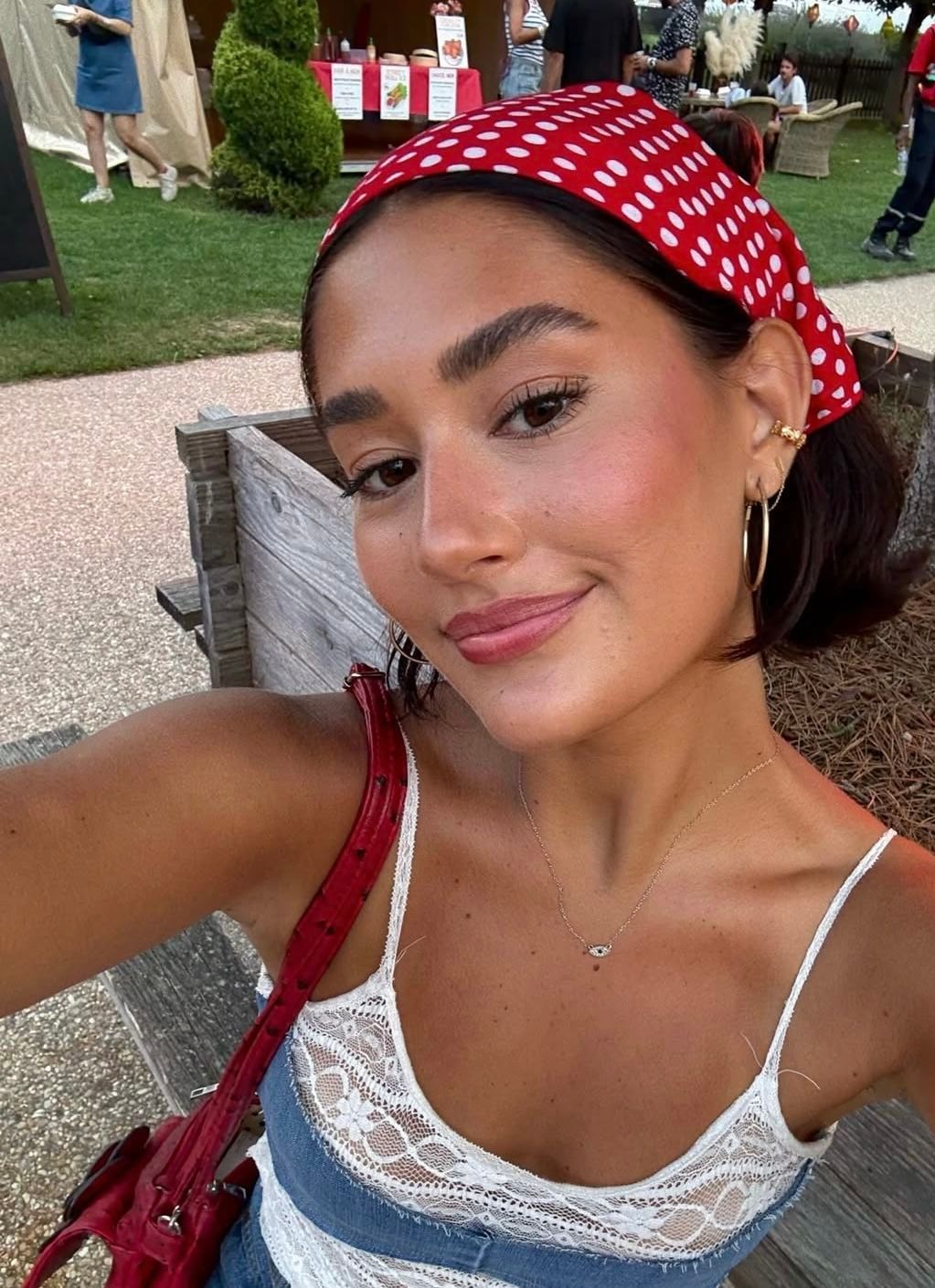Young woman wearing red polka dot headband and white lace top takes outdoor selfie with red handbag.