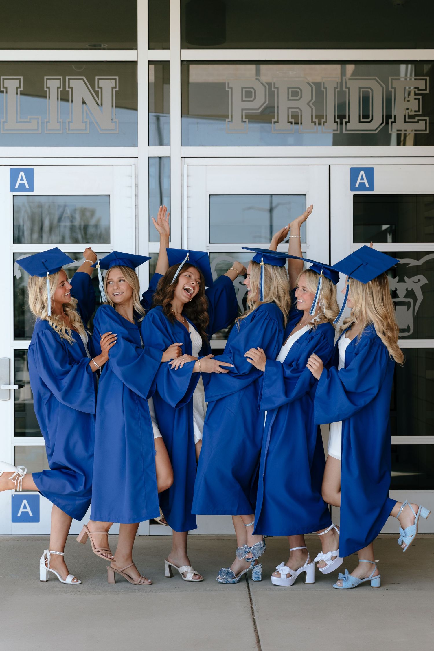Group of graduates in blue caps and gowns pose together against a building wall during graduation ceremony.