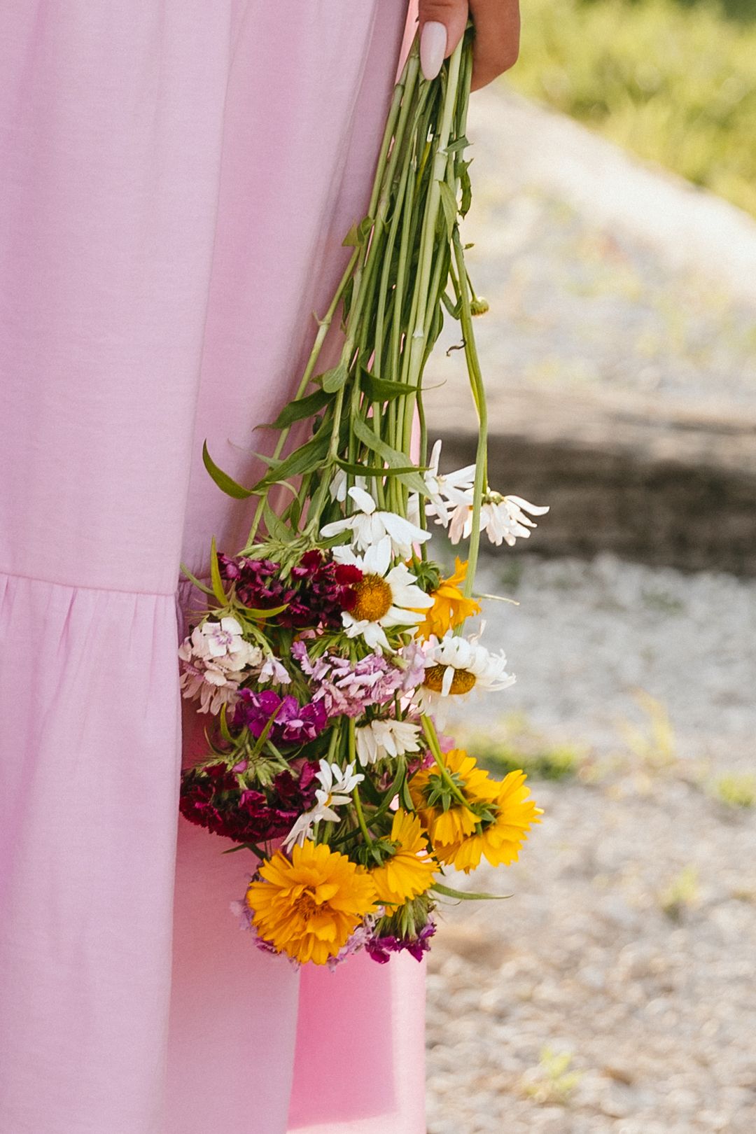 Person in pink dress holding a colorful wildflower bouquet with daisies and marigolds outdoors.