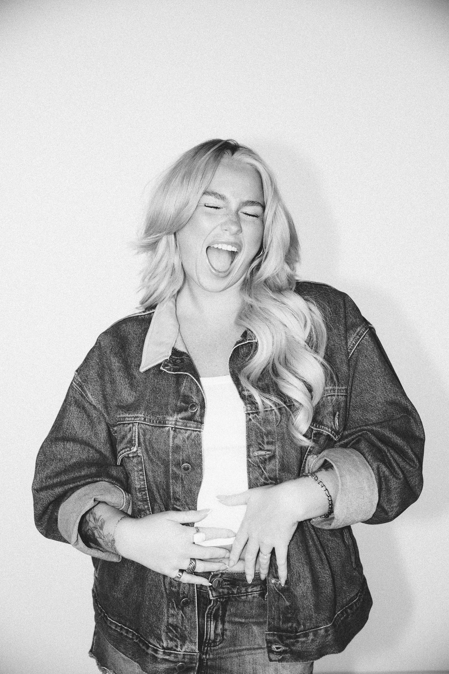 Laughing blonde woman in oversized denim jacket and white tee, black and white photo.