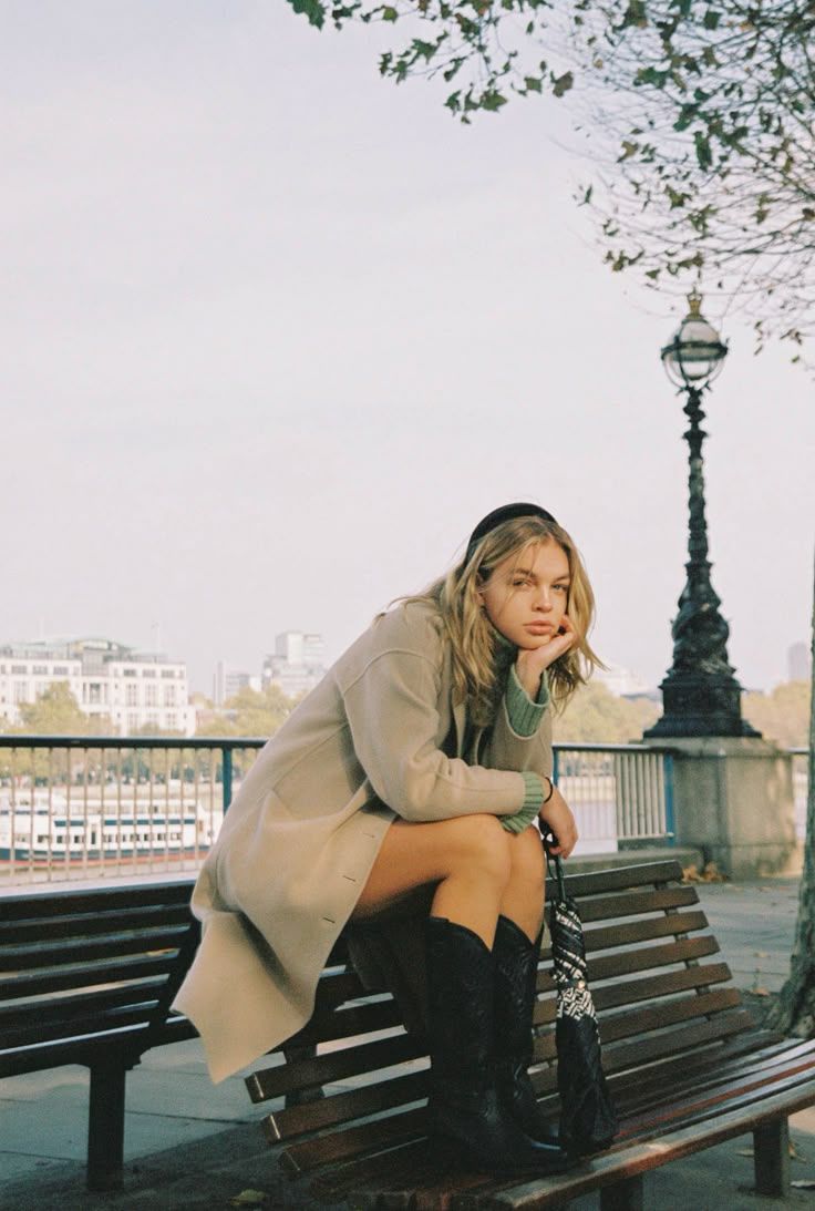 Woman in a beige coat and black knee-high boots sits on a wooden bench by a riverside railing near a vintage lamppost.