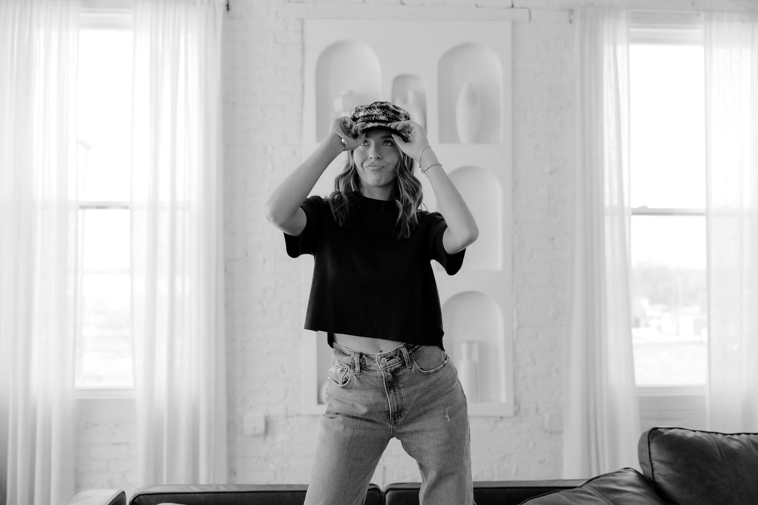 Black and white photo of a woman adjusting a patterned hat in a bright, modern living room.