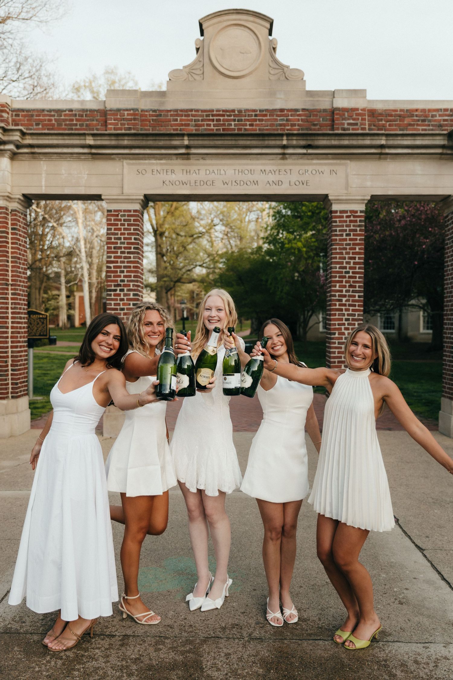 Five women in white dresses celebrating graduation, toasting with champagne bottles in front of a brick university arch.