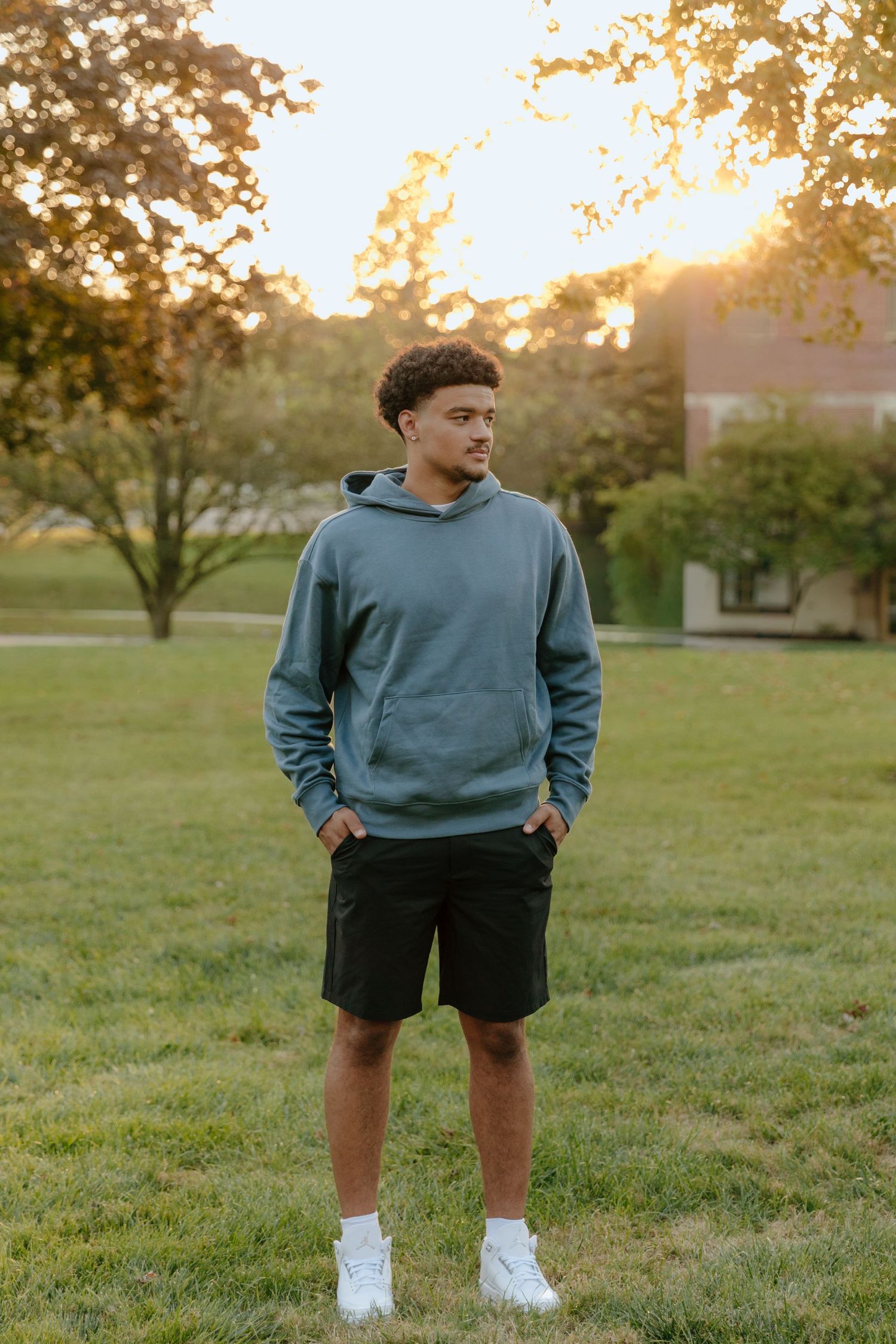 Young man in teal hoodie and black shorts stands on grass during golden hour sunset in park.