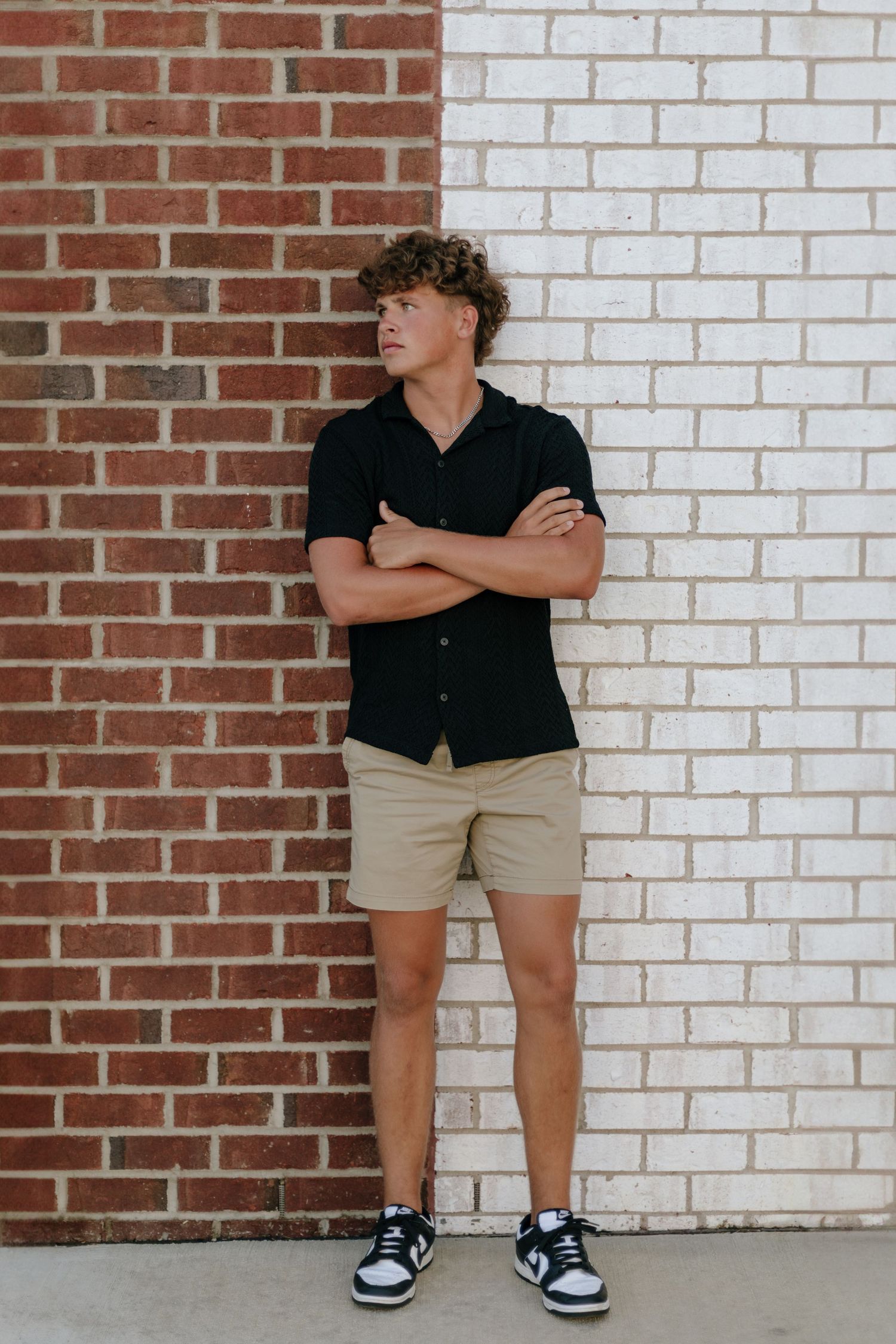 Person in casual summer attire with black shirt and khaki shorts poses against two-toned brick wall.